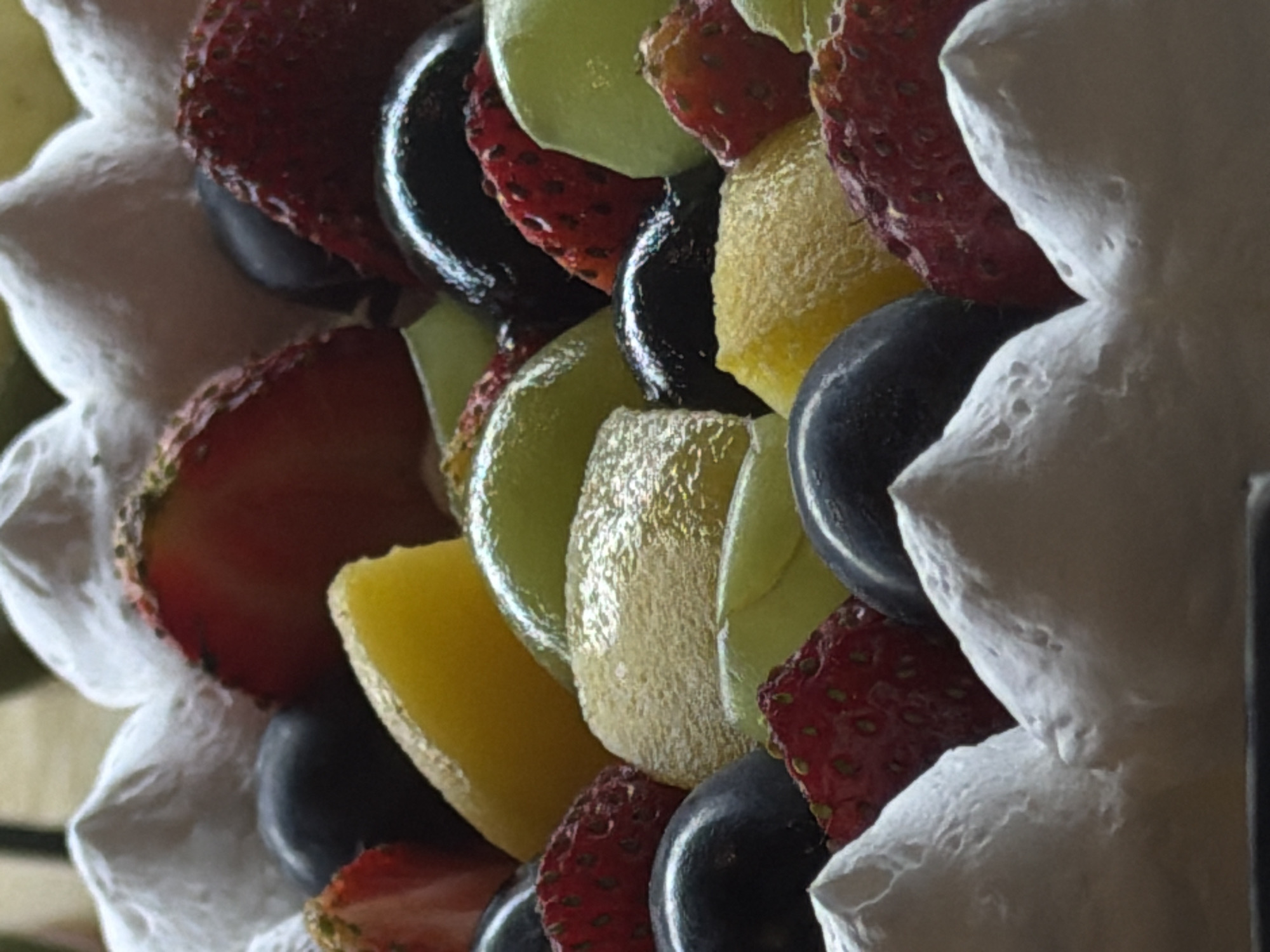 The image presents a close-up, vertical-oriented view of a vibrant dessert or fruit arrangement, likely taken in North Kuta, Indonesia, based on the provided context. The primary subjects are an assortment of fresh fruits and dollops of white, textured cream or frosting.
The fruits visible include bright red, sliced strawberries with visible seeds, dark, glossy black grapes, light green melon slices (possibly honeydew), and yellowish fruit slices, which could be mango or pineapple. These fruit pieces are arranged in a dense, layered pattern, interspersed with generous swirls of white cream, which has a fluffy, peaked texture. The arrangement suggests a decorative presentation, possibly part of a cake, tart, or fruit platter.
No human activity is directly depicted, but the careful arrangement of the ingredients implies the prior activity of food preparation. The lighting is soft, highlighting the natural sheen and moisture on the fruit surfaces. There are no specific location cues within the image itself, nor are there any indications of time of day, weather, or visible text. The focus is entirely on the appetizing textures and contrasting colors of the fruit and cream, creating an inviting visual.