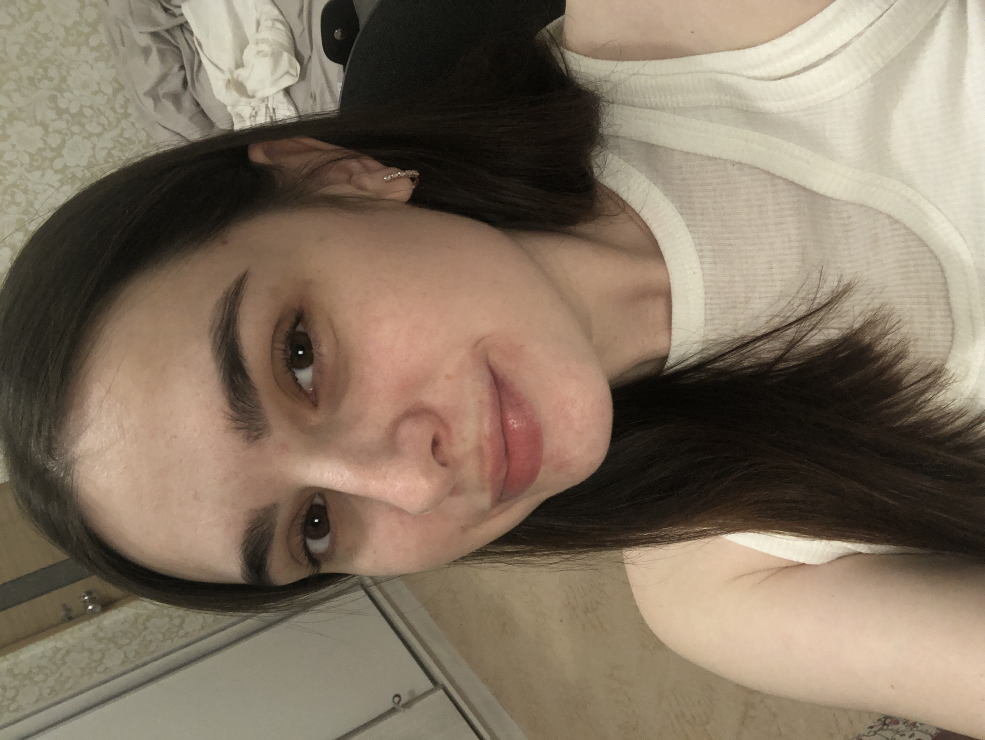 A young woman from Noviy, Russia, is captured in a selfie-style photograph, rotated 90 degrees clockwise. She is shown from the chest up, with long, dark brown hair that frames her face. Her dark eyebrows are well-defined, complementing her dark eyes. She has a soft, slight smile on her lips, which are naturally pink with a hint of shine. She is wearing a plain white ribbed tank top. A small, sparkling earring is visible in her right ear. Her skin appears natural with some subtle redness. The background suggests an indoor setting, possibly a bedroom. To her left, a light-colored wall features a subtle patterned wallpaper. Behind her head, rumpled clothes or fabric in shades of white and grey, along with a dark object (possibly part of furniture), are visible. The lighting is soft, indicating an indoor environment. Her expression is calm and relaxed. No text is visible in the image.