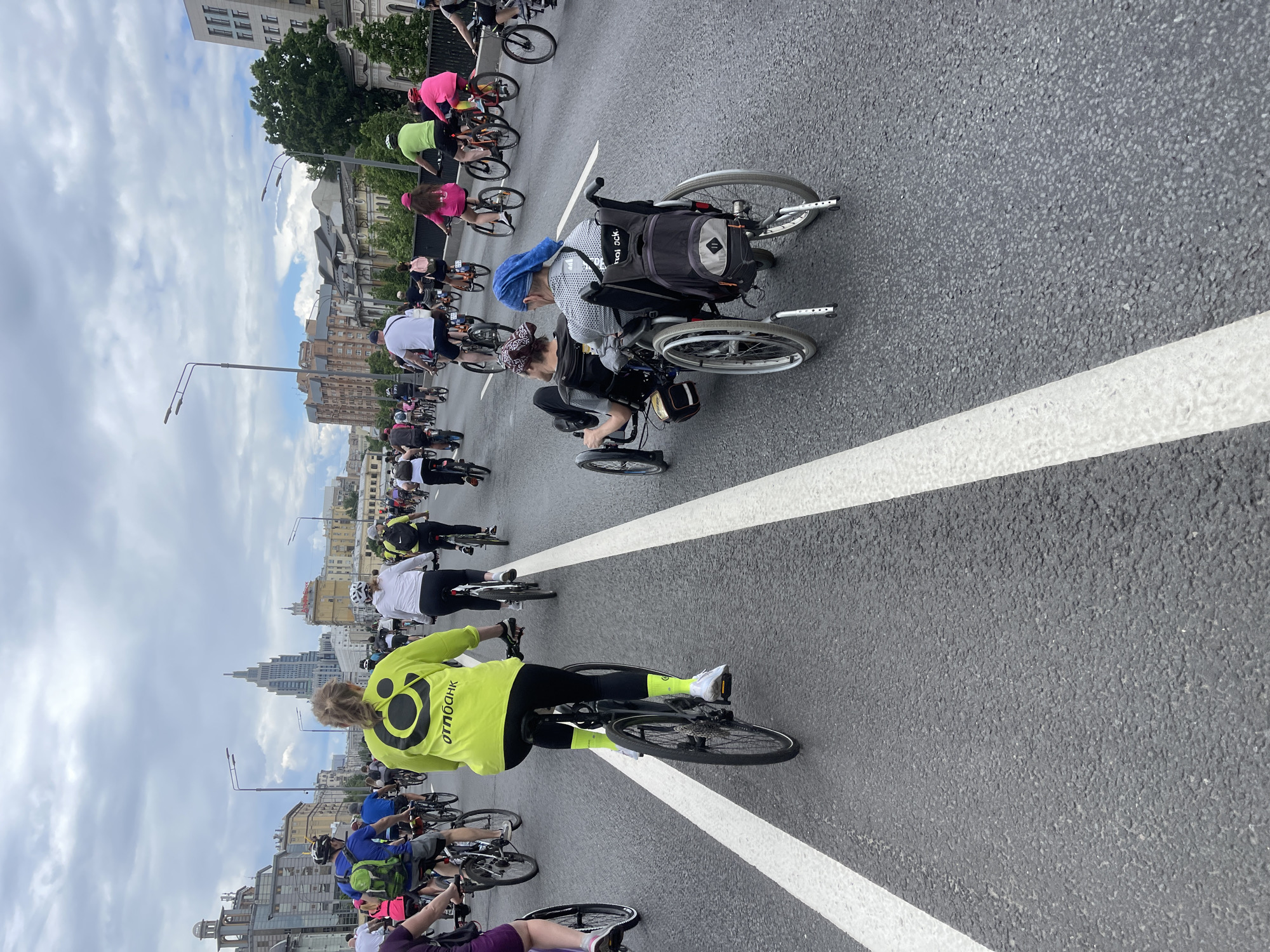 The image shows a bicycle parade in Moscow, Russia, on a wide asphalt road marked with thick white dividing lines. Many people are participating, most riding bicycles. Some individuals are using specialized hand-powered bicycles or wheelchairs. The participants are dressed in varied athletic attire, with some wearing bright neon colors. Buildings and streetlights line the background. The sky is overcast with cloudy weather.