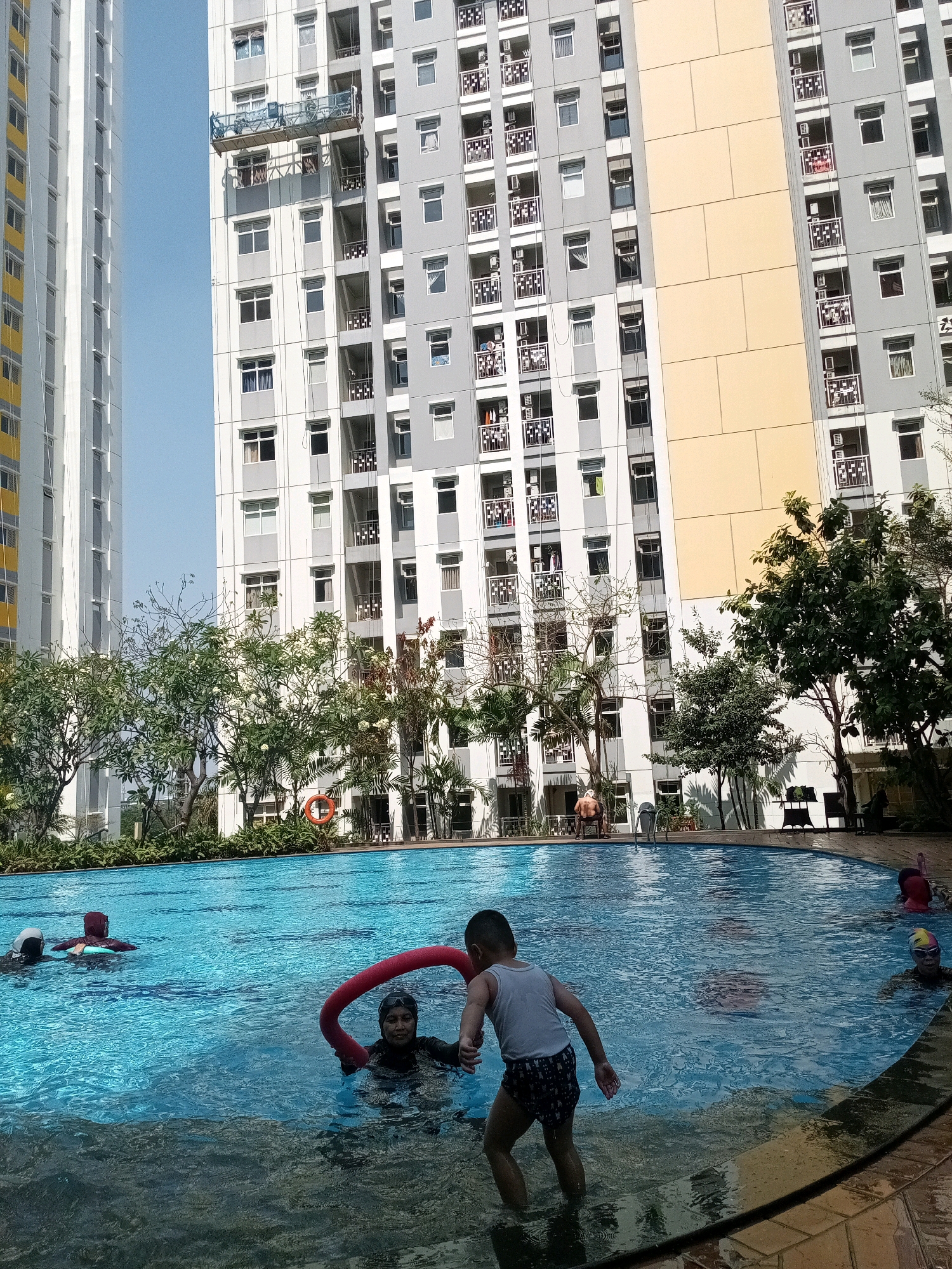 The image depicts a scene at a swimming pool in Bekasi, Indonesia, likely within a residential complex. In the foreground, a young boy in a white tank top and patterned swim trunks stands at the edge of the pool, facing away from the camera. To his left, a woman wearing a dark swimsuit and swim cap is partially submerged in the water, holding a red pool noodle. In the middle ground, several other individuals are visible in the blue water, including a person wearing a red swim cap and another with a dark swim cap. The pool area is surrounded by lush green trees and foliage, with tall, modern apartment buildings in the background. One building is yellow and cream-colored, while another is predominantly gray and white. The sky is clear and blue, suggesting a sunny day. A life preserver ring is visible on the side of the pool.