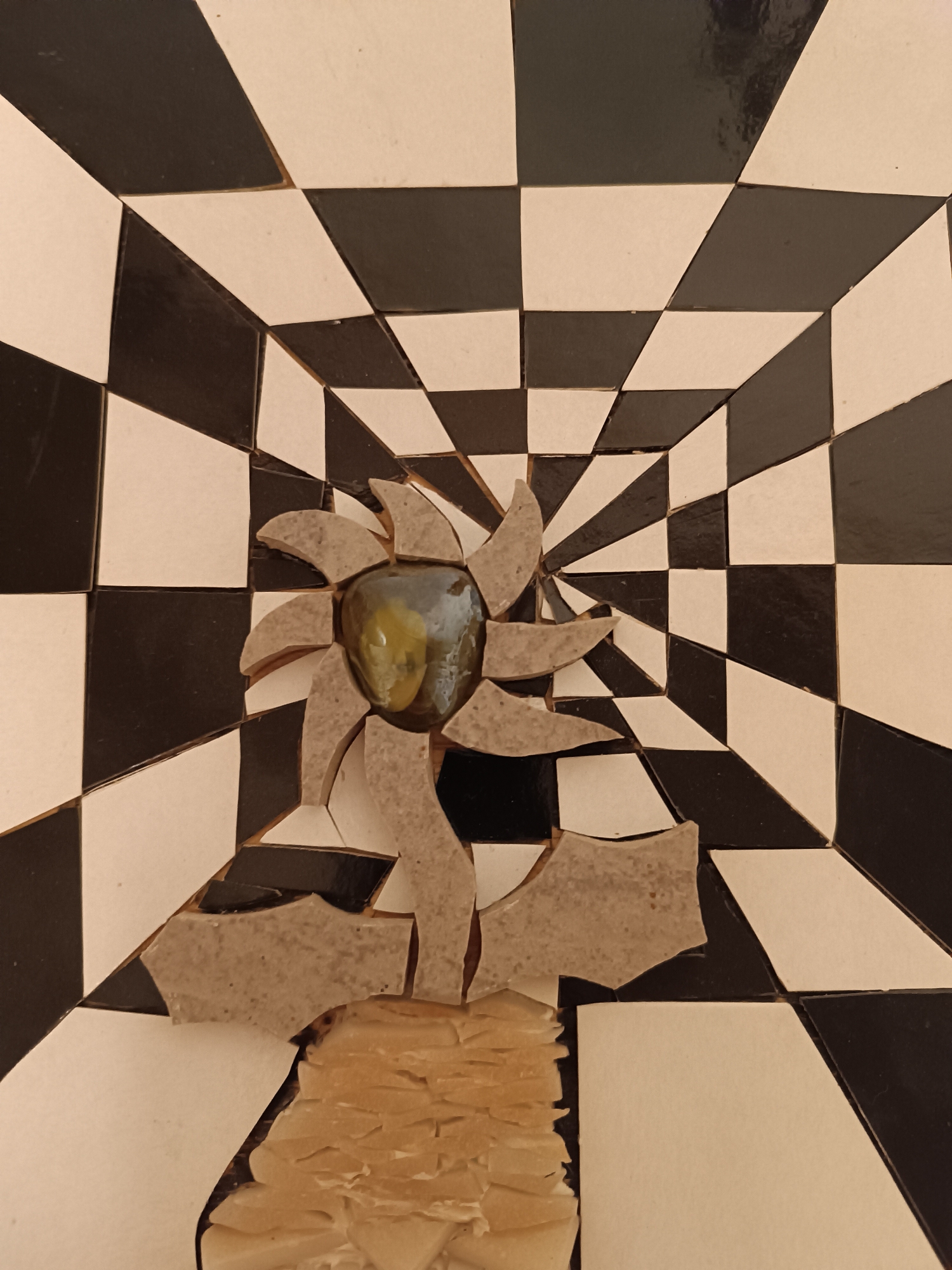 This image captures a mosaic artwork, likely from Viadana, Italy, characterized by an abstract design and a strong optical illusion in the background.
The background features a dynamic checkerboard pattern of black and off-white squares. These squares are distorted and arranged to create a powerful sense of receding depth, drawing the viewer's eye towards the center as if looking down a tunnel or into a warped space.
In the foreground, centrally positioned, is a mosaic representation that evokes a stylized flower or sun. Its core is a prominent, rounded, greenish-yellow piece, possibly translucent glass or a polished stone, which reflects light. Surrounding this central element are irregularly shaped, light brown or beige mosaic tiles, arranged like petals or rays emanating outwards. A stem-like structure, composed of similar light brown/beige mosaic pieces, extends downwards from the main figure. The base of the artwork, beneath the stem, is formed by many smaller, irregular, pale yellow or cream-colored mosaic shards, layered and stacked to create a textured, organic appearance, possibly representing roots or ground.
The artwork utilizes a muted, earthy color palette and incorporates hand-cut or broken tiles, giving it an artisanal and somewhat rustic quality. There is no discernible text within the image.