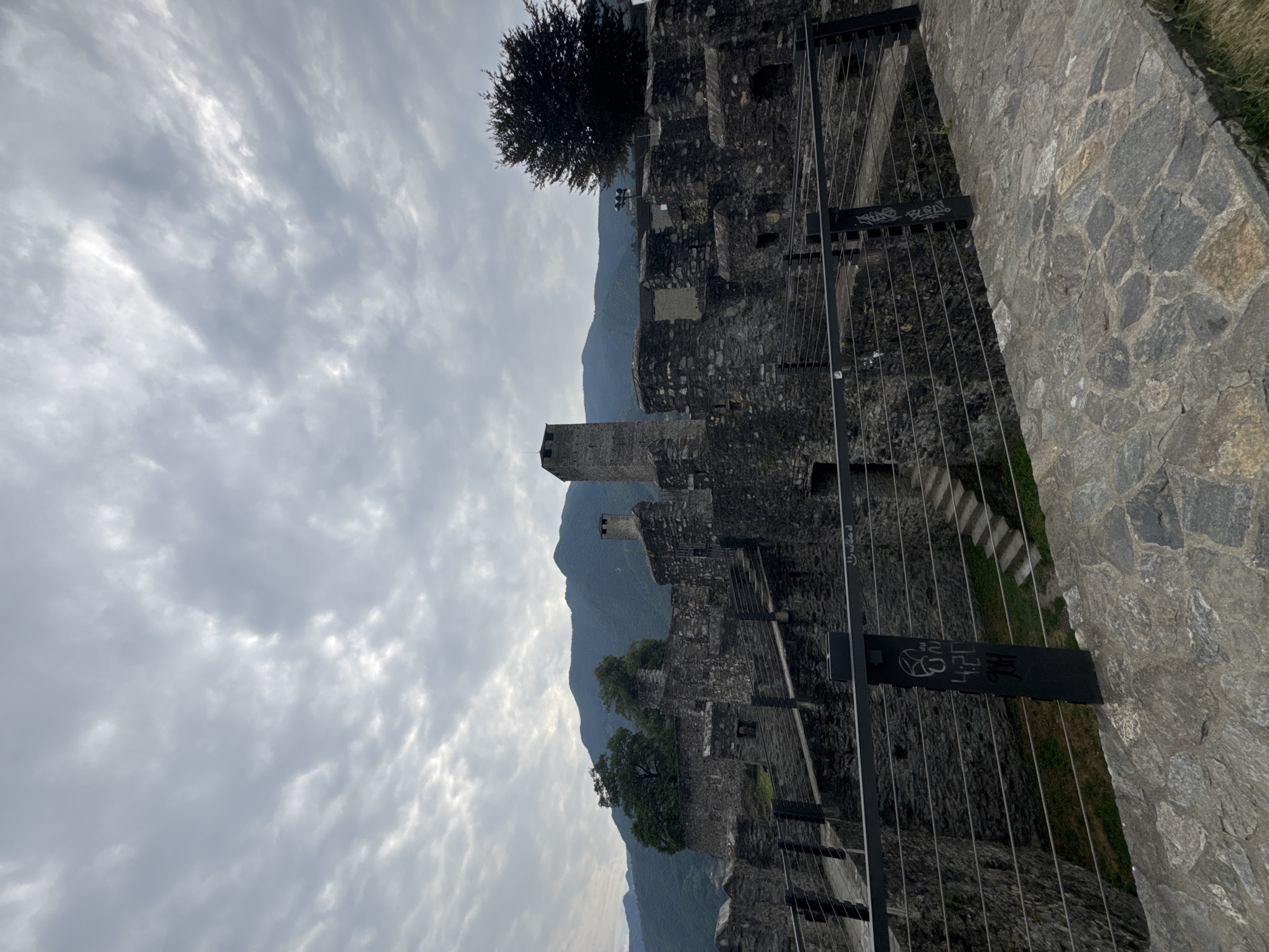 The image shows a view of the Castel Grande in Bellinzona, Switzerland, under a cloudy sky. The castle is constructed of dark stone and features walls, towers, and battlements. A pathway paved with irregular stones leads the viewer's eye towards the castle. A black metal railing with thin wire cables runs along the edge of the pathway. Beyond the railing, stone stairs ascend within the castle grounds, and more stone walls and structures are visible. In the background, lush green mountains rise against the overcast sky, with a dark-leaved tree to the right of the castle. The castle appears to be a historical site, with some signs of age and wear. There are no people visible in the image.