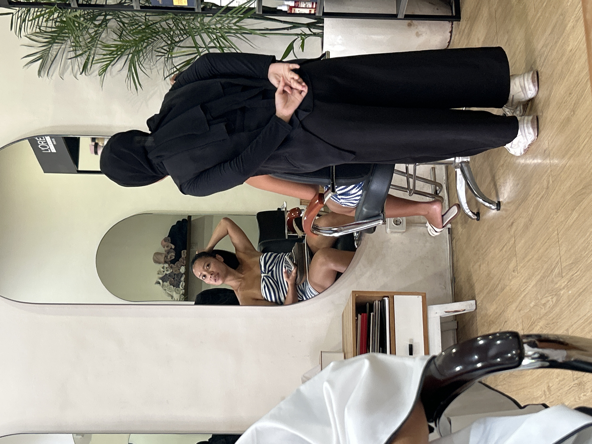 In a salon in North Kuta, Indonesia, a woman wearing a black hijab and black wide-leg pants stands with her hands clasped behind her back. She is facing away from the camera, towards a mirror. In the reflection of the mirror, a woman in a blue and white striped tube top is seated, touching her hair and holding a phone and a small brown object. She is positioned in a salon chair. A white fabric is draped over a chair in the foreground, obscuring part of the scene. A wooden shelf unit with drawers is visible to the left of the mirrored area. A tall potted plant stands to the right of the woman in the hijab. The flooring is light-colored wood.