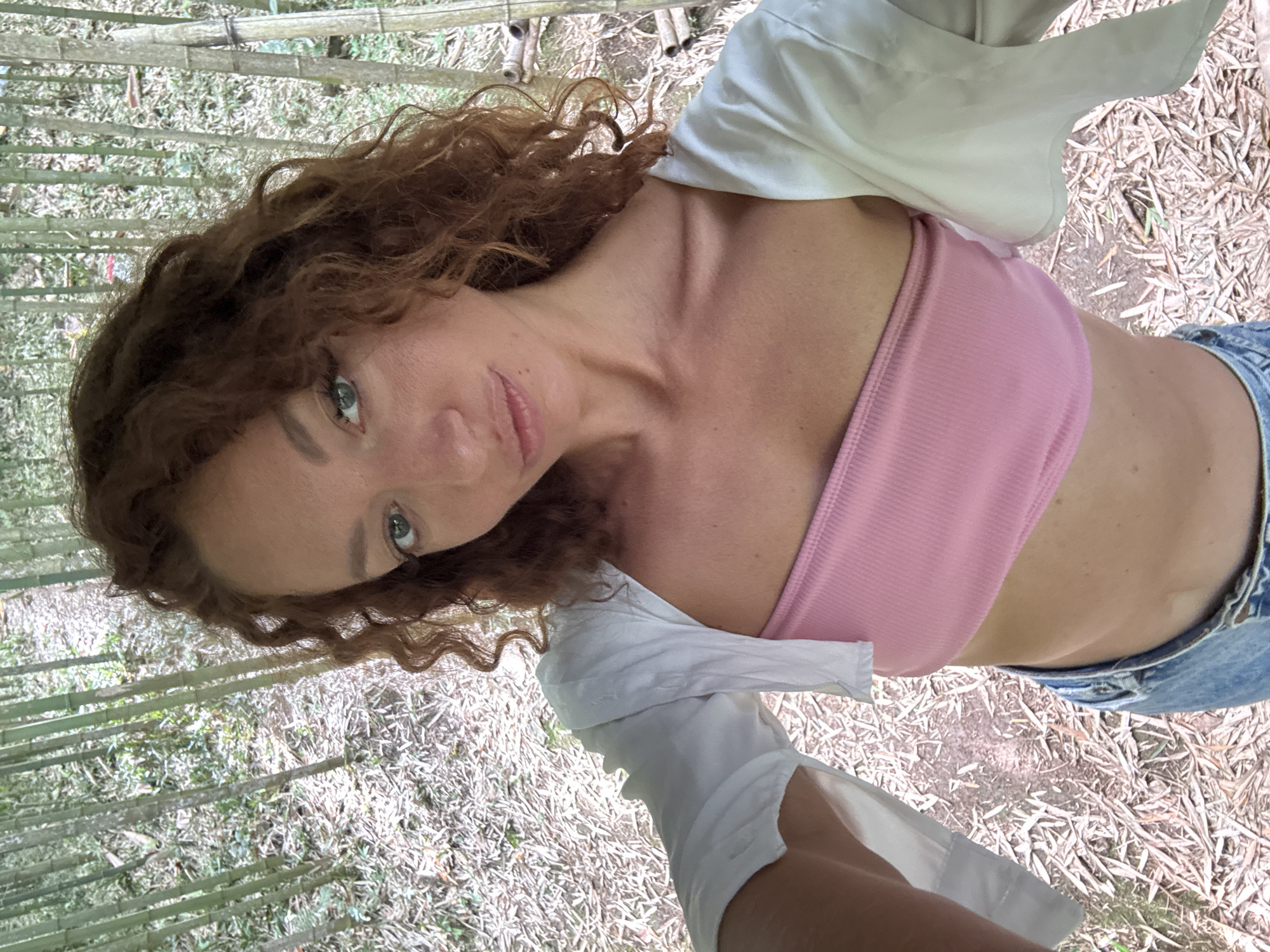 A woman is standing in a bamboo grove in Ngã Ba Kim, Vietnam. She is wearing a pink top, blue jeans, and a white shirt. The woman is looking at the camera. The background is a bamboo forest.