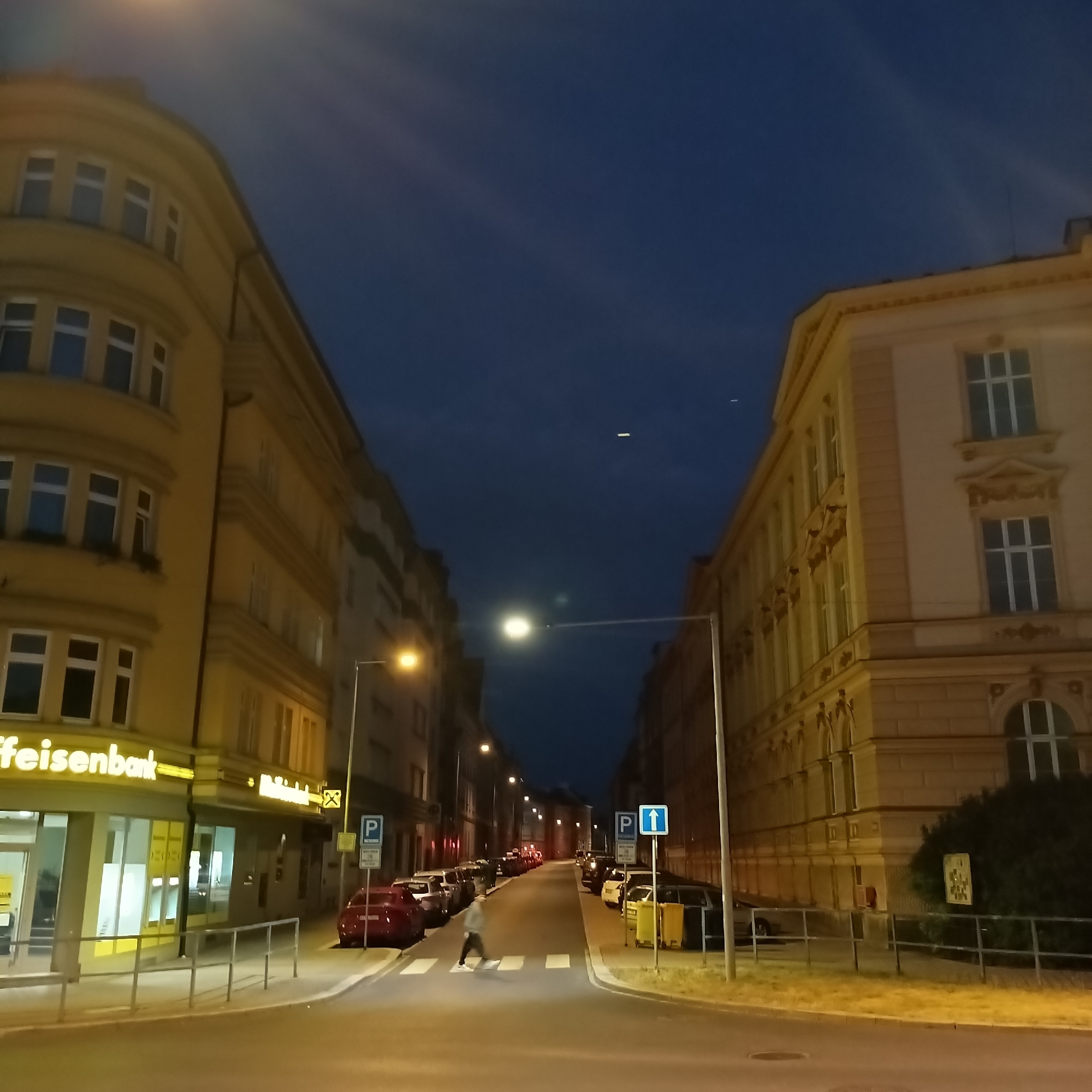 The image depicts a street scene at dusk in Cheb, Czechia. On the left, a yellow building with a rounded corner features multiple windows. The ground floor of this building has large glass windows, one displaying signage that reads "raiffeisenbank". A metal railing runs along the sidewalk in front of the building.
On the right, a lighter colored building with classical architectural elements, including ornate window frames and cornices, is visible. This building also has numerous windows. Between the buildings, the street is lined with parked cars. A red car is parked on the left side of the street.
A crosswalk is marked on the asphalt road, and a person wearing a light-colored hooded jacket is crossing it, moving from right to left in the frame. Streetlights illuminate the scene, casting a warm glow. Traffic signs, including a blue parking sign with a white "P", are present on the right side of the street. The sky is a deep blue, indicating the transition from day to night, with some faint wispy clouds.