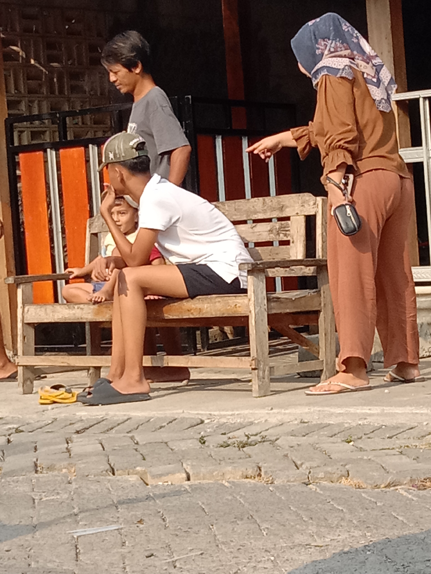 A man in a grey t-shirt stands and looks down to his left. To his right, a woman wearing a brown headscarf and brown outfit points to her left, towards a wooden bench. Seated on the bench are two individuals. The person in the foreground, wearing a white t-shirt and black shorts, looks to their left and has their hand raised near their mouth. To their left, a younger individual in a yellow shirt sits with their arms crossed. A pair of grey slippers and a yellow sandal are on the cobblestone ground in front of the bench. In the background, a fence with orange and white slats is visible, along with parts of a building. The setting appears to be outdoors, likely in a public area within the city of Wirosari, Indonesia, given the architectural style and the cobblestone pavement.