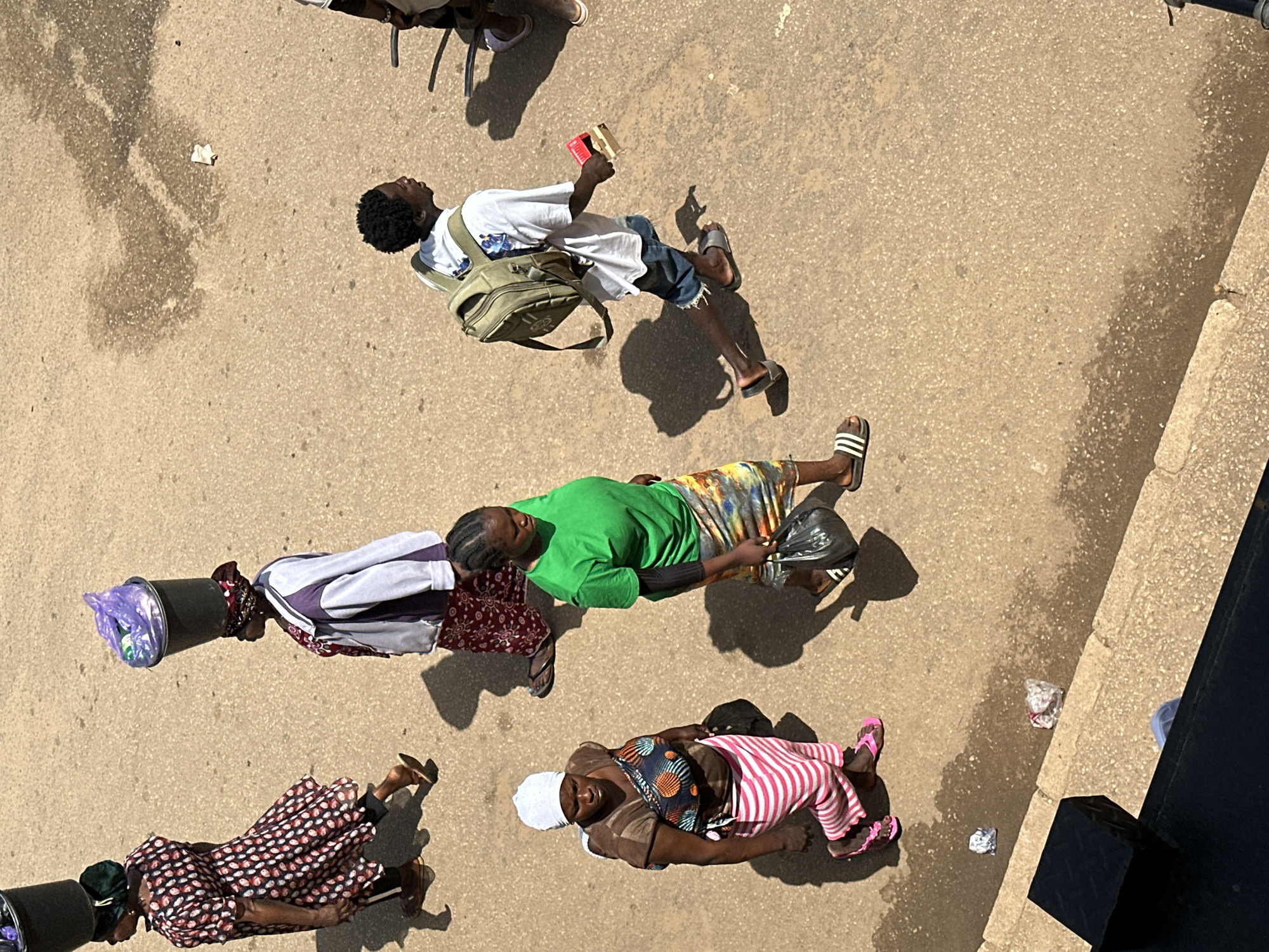 In Bukuru, Nigeria, five individuals walk across a dusty street under bright sunlight, casting distinct shadows. A woman on the far left carries a large black bucket on her head, wearing a patterned dress and sandals. Next to her, another woman balances a similar black bucket filled with a purple plastic bag. She wears a patterned skirt and a white jacket over a red garment. In the center, a woman in a green t-shirt and a colorful, patterned skirt walks with a black plastic bag in her right hand. To her right, a young man with a backpack walks while holding a red object in his right hand and wearing denim shorts and sandals. The southernmost person visible, a woman wearing a white headscarf, a brown shirt, and a pink and white striped skirt, is seen from the waist down, carrying a dark bag and wearing pink sandals. Scattered across the ground are small, reflective pieces of litter. The road surface is a mix of asphalt and gravel, with some damp patches.