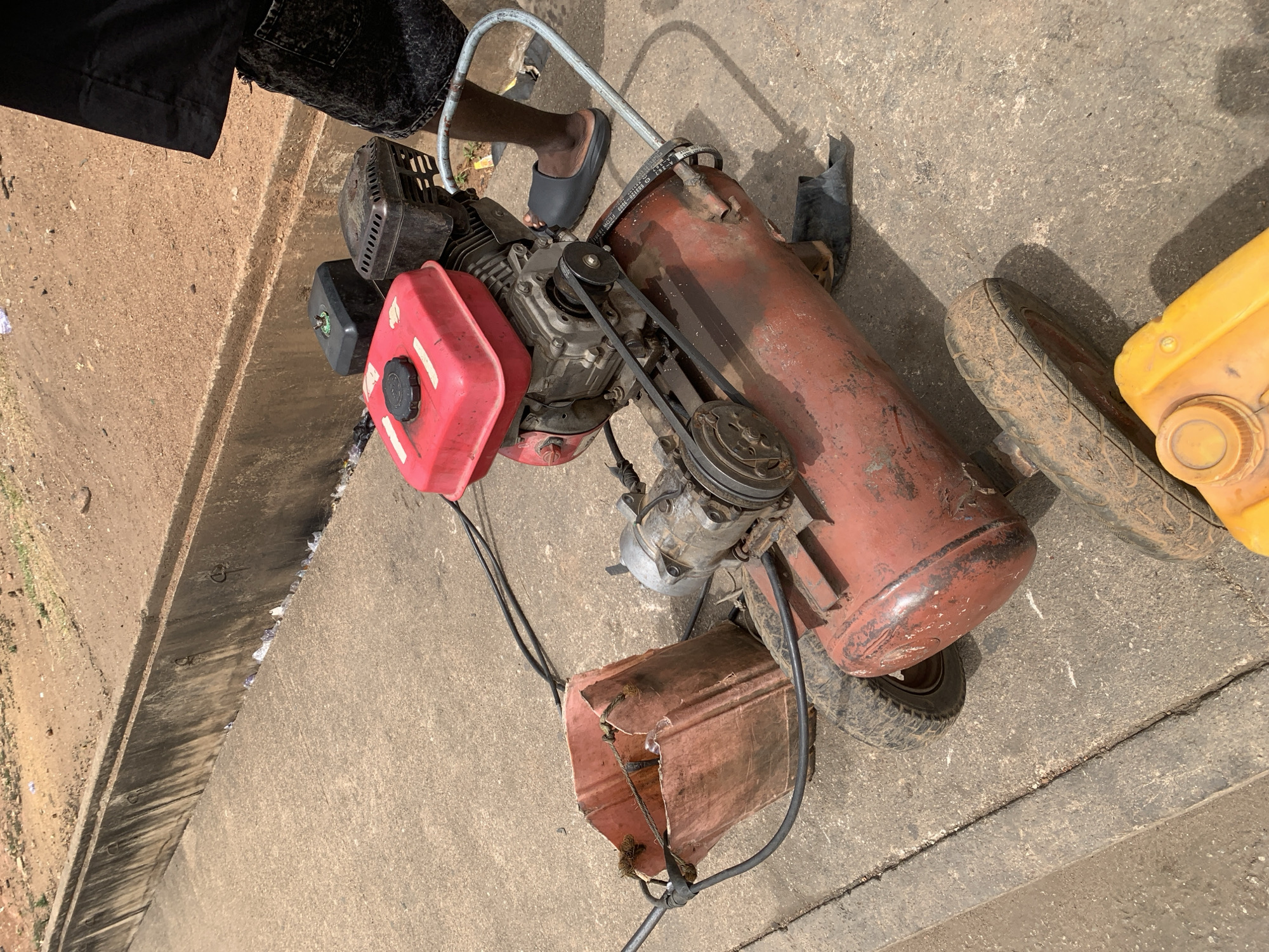 A worn-looking, red air compressor sits on a concrete surface. The compressor consists of a motor with a red fuel tank, connected via belts to a cylindrical tank. The compressor has wheels. A person wearing a dark-colored garment and black footwear stands near the compressor. To the side, there is a partially visible yellow container. The location appears to be an outdoor space in Jos, Nigeria, based on the style and architecture of the surrounding infrastructure and typical road markings.