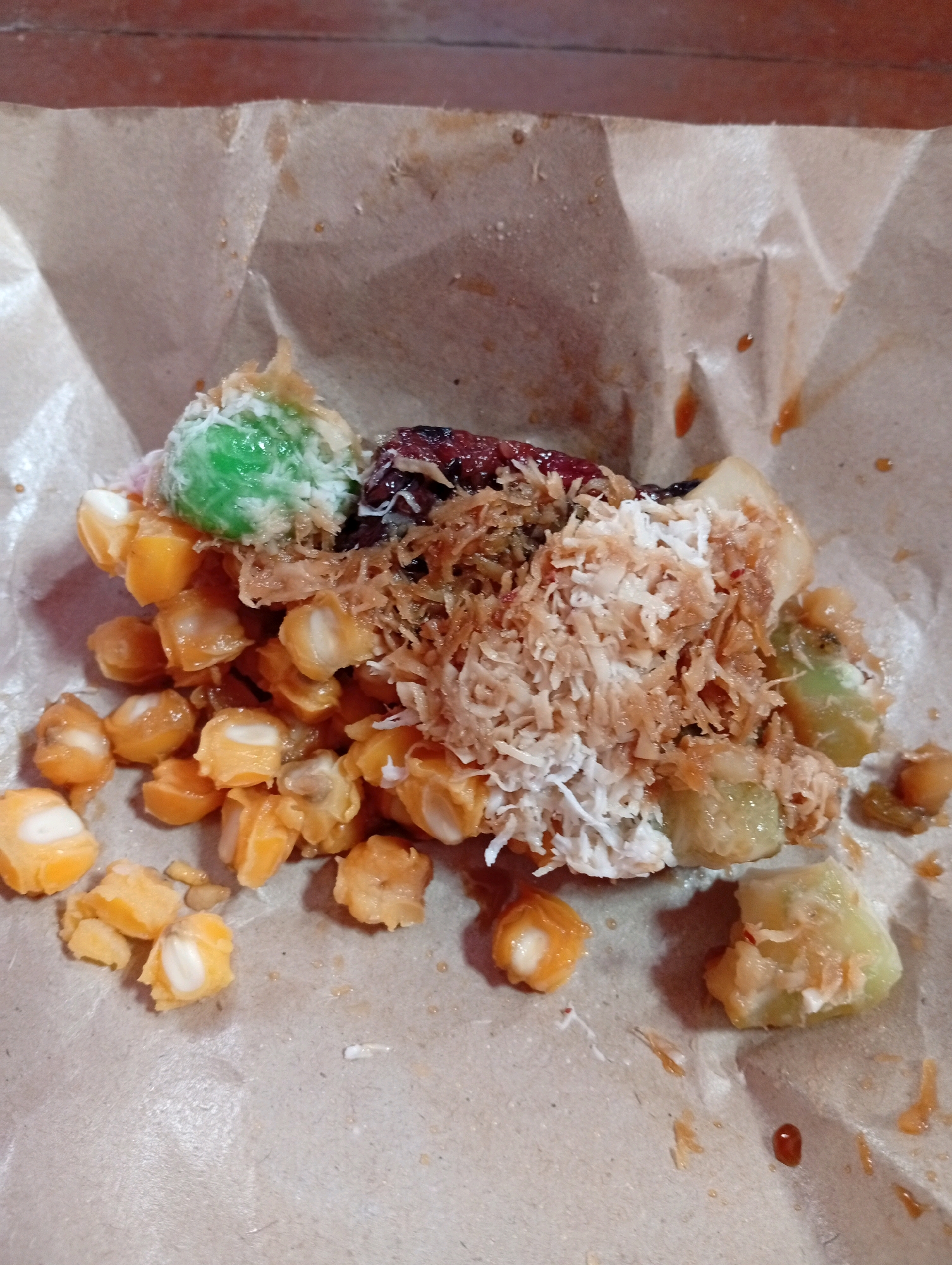 A close-up, overhead shot captures a pile of Indonesian street food resting on brown paper. The dish features a base of sweet corn kernels, interspersed with pieces of green pandan-flavored glutinous rice balls coated in shredded coconut. A dark, jam-like topping, possibly palm sugar syrup, is visible amongst the coconut. The food appears to be served as a single portion. The brown paper wrapper is crumpled and shows some moisture stains, suggesting it may be a takeaway item. The background is blurred, hinting at a rustic, outdoor setting, common for street food vendors in cities like Salatiga, Indonesia. There are no people or animals visible in the image.