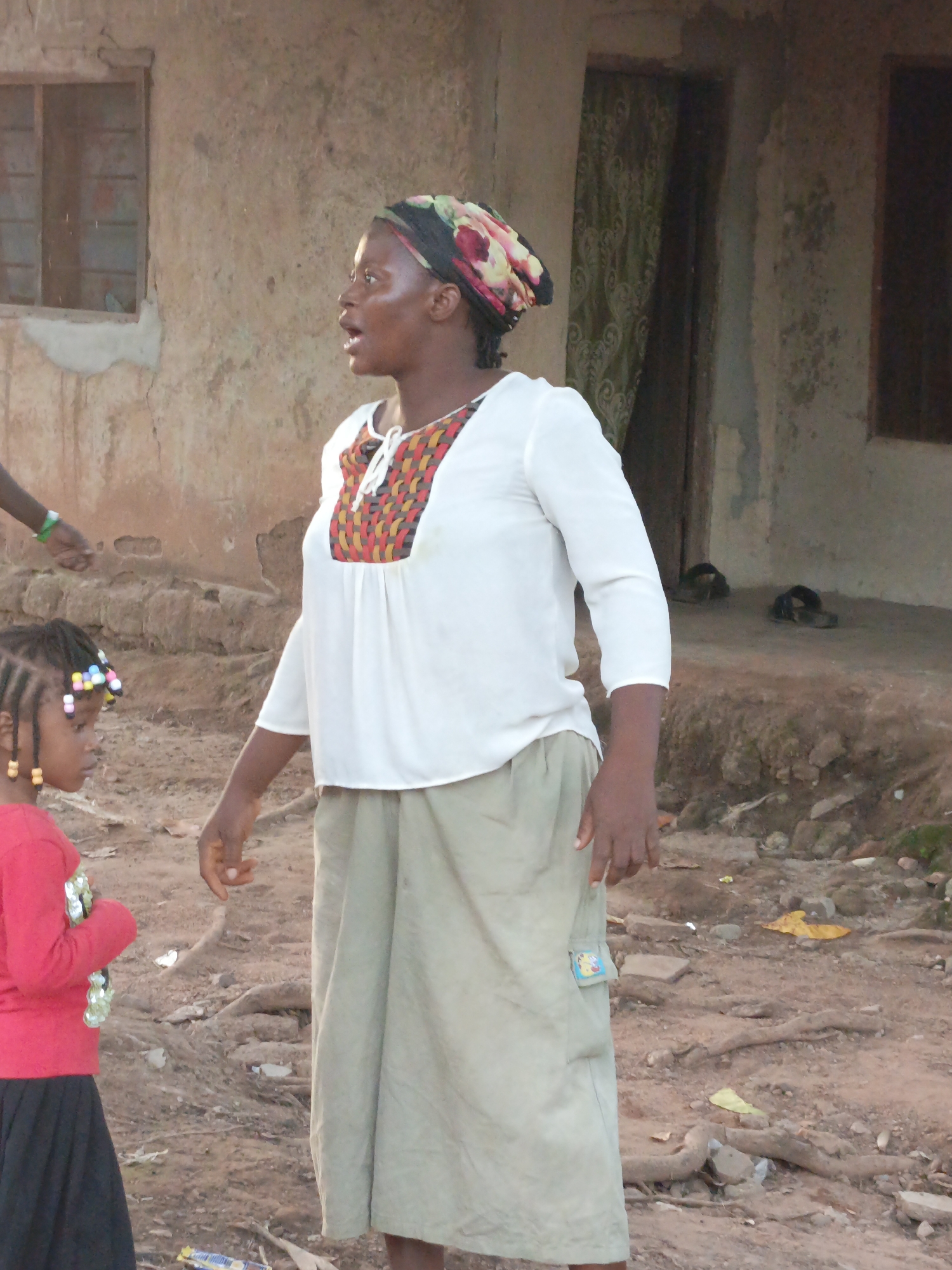 In Akwanga, Nigeria, a woman stands outside a weathered building with a small window. She wears a colorful headwrap and a white blouse with a patterned yoke. A young girl, her hair adorned with beads, stands to her left, wearing a red long-sleeved shirt and a black skirt. The ground around them is dirt with scattered debris, including some yellow leaves and what appears to be a discarded wrapper. In the background, a doorway with a patterned curtain is visible, and a pair of sandals rests on the ground near the entrance.