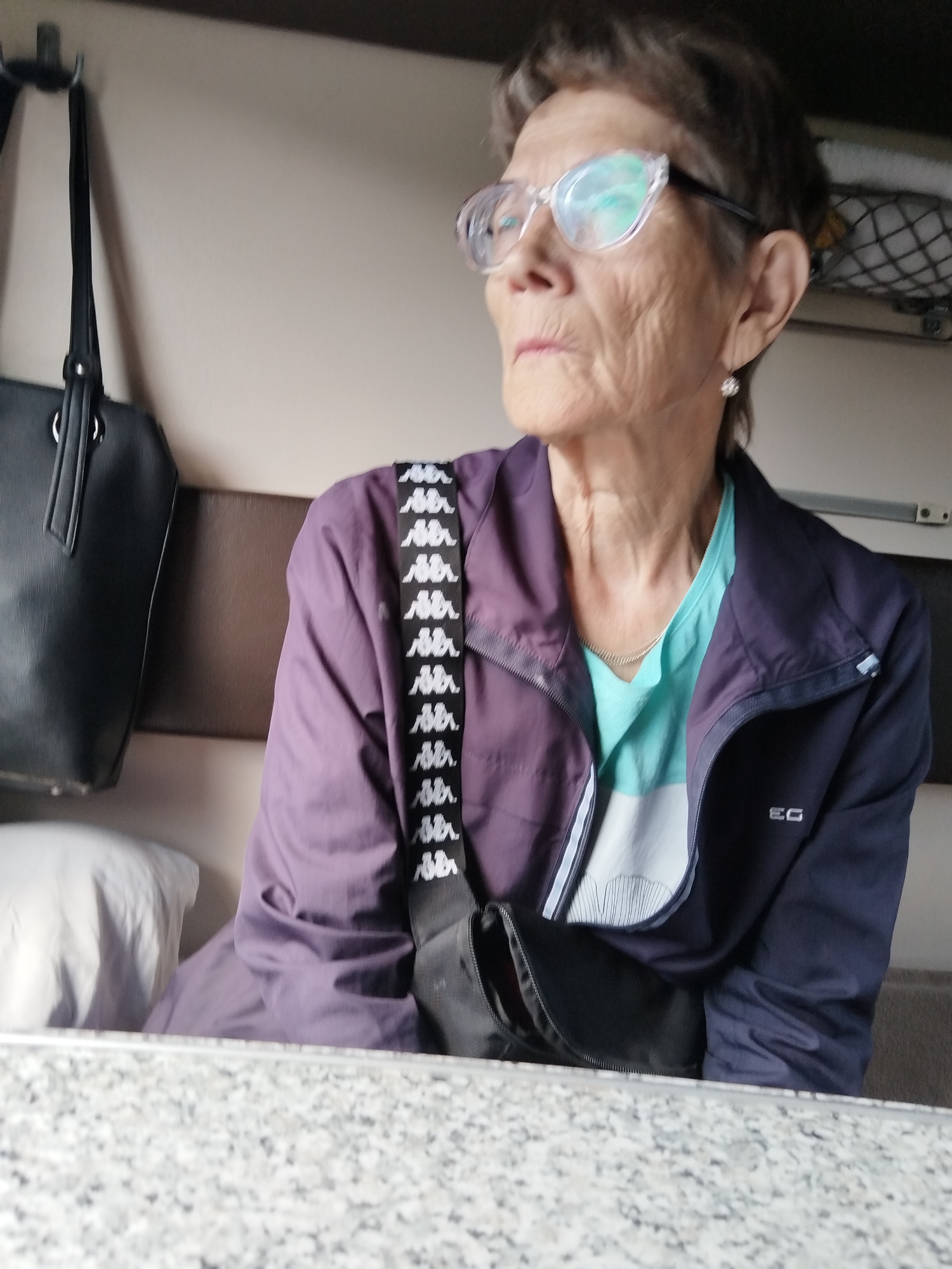 An elderly woman with short brown hair and clear-rimmed glasses is seated, looking to her left. She wears a purple jacket over a turquoise shirt. A black crossbody bag with a white logo pattern hangs from her shoulder. To her left, a large black tote bag is visible. The background suggests an interior setting, possibly a train car, with a brown headrest and a light-colored wall. A white pillow is on her left. The foreground shows a speckled gray surface. The image is dated 2798 Km, Russia.