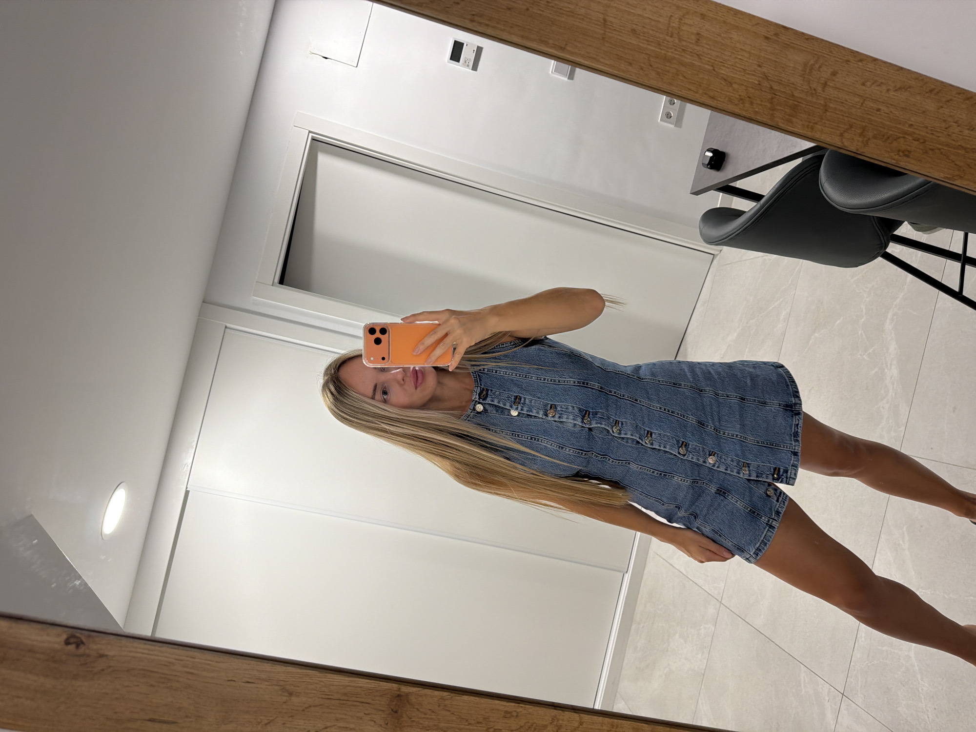 A woman with long blonde hair is taking a mirror selfie in what appears to be a modern apartment or hallway. She is wearing a blue denim dress with buttons down the front. She is holding an orange smartphone up to take the photo, and her reflection is visible in a large mirror that takes up most of the frame. The background of the reflection shows a white wall, a light-colored tiled floor, and a wooden frame around the mirror. In the reflection, there is also a glimpse of some furniture, including a black chair and a table, and some light fixtures on the wall. The lighting suggests it is daytime. The overall impression is one of casual elegance. The location context suggests this is in Finestrat, Spain.
