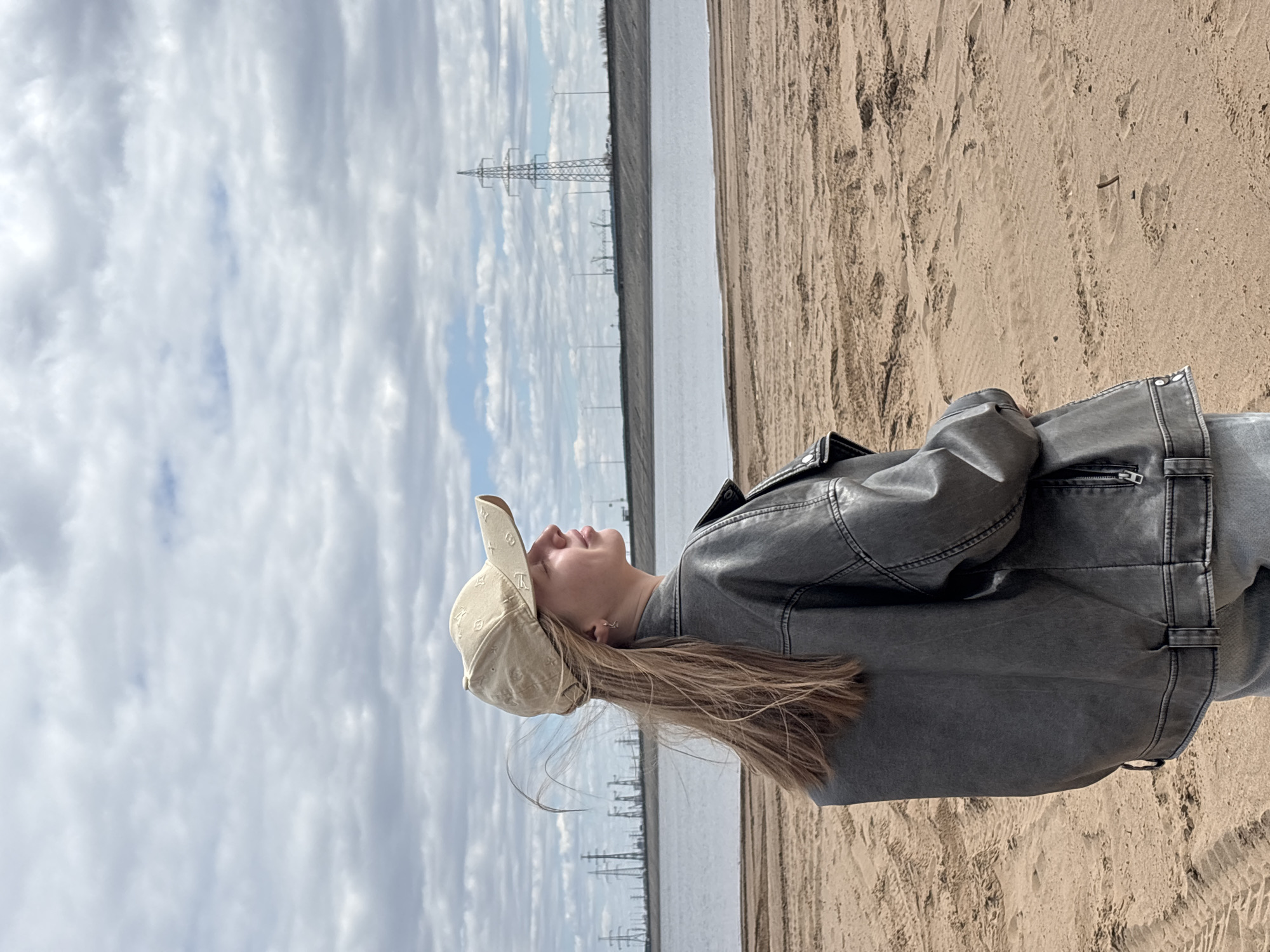 A young person wearing a baseball cap and jacket stands in a sandy area next to a body of water. The background includes a cloudy sky and a series of electricity pylons. The location is Noviy, Russia.