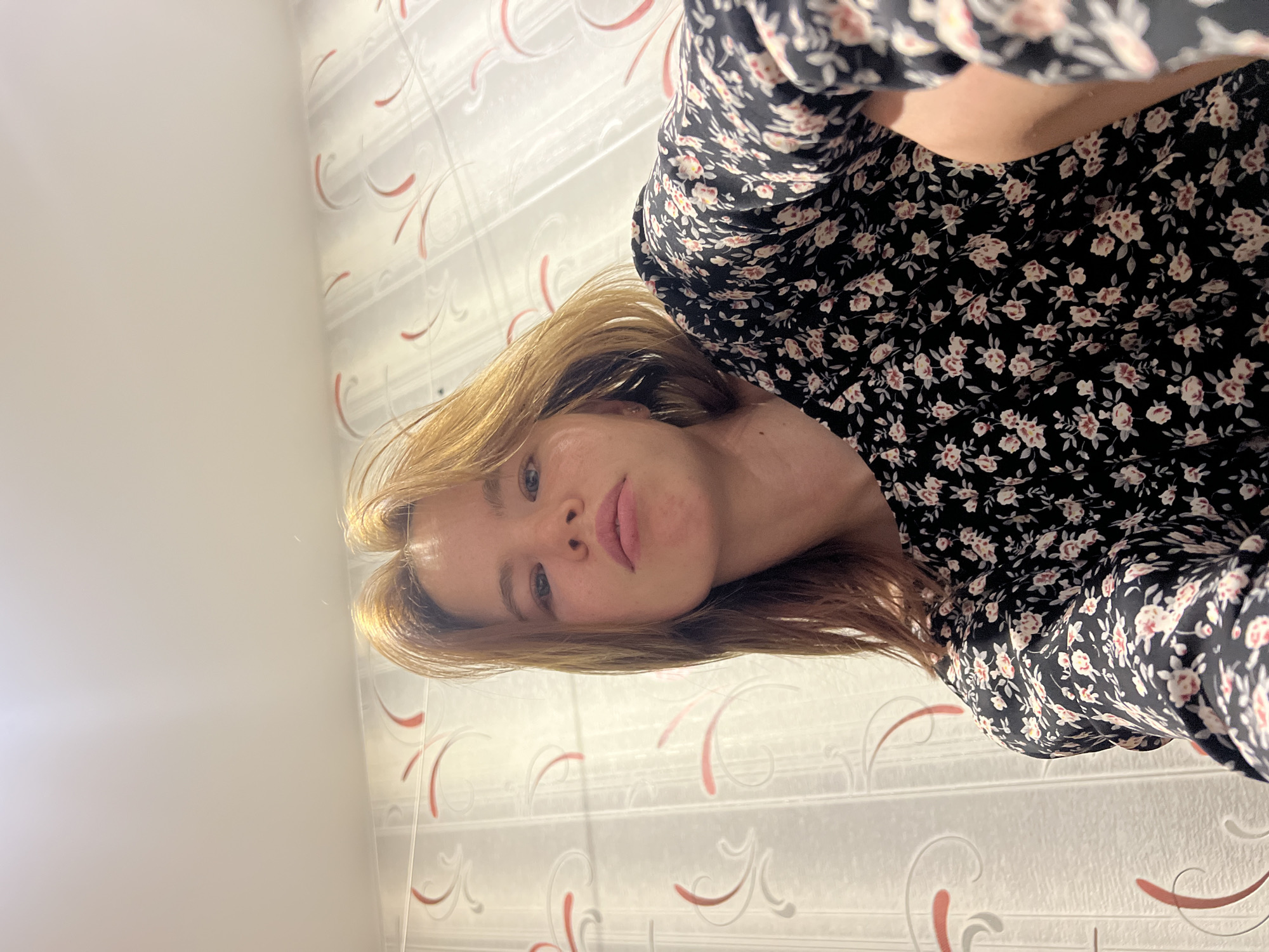 A young woman with blonde hair and light skin is looking into the camera with a neutral expression. She is wearing a black dress with a floral print. The background appears to be a wall with patterned wallpaper. The image is taken from a low angle, suggesting the photographer is positioned below the subject. There are no specific identifiable landmarks or clues that would place this image in the city of Chaikovsky, Russia.