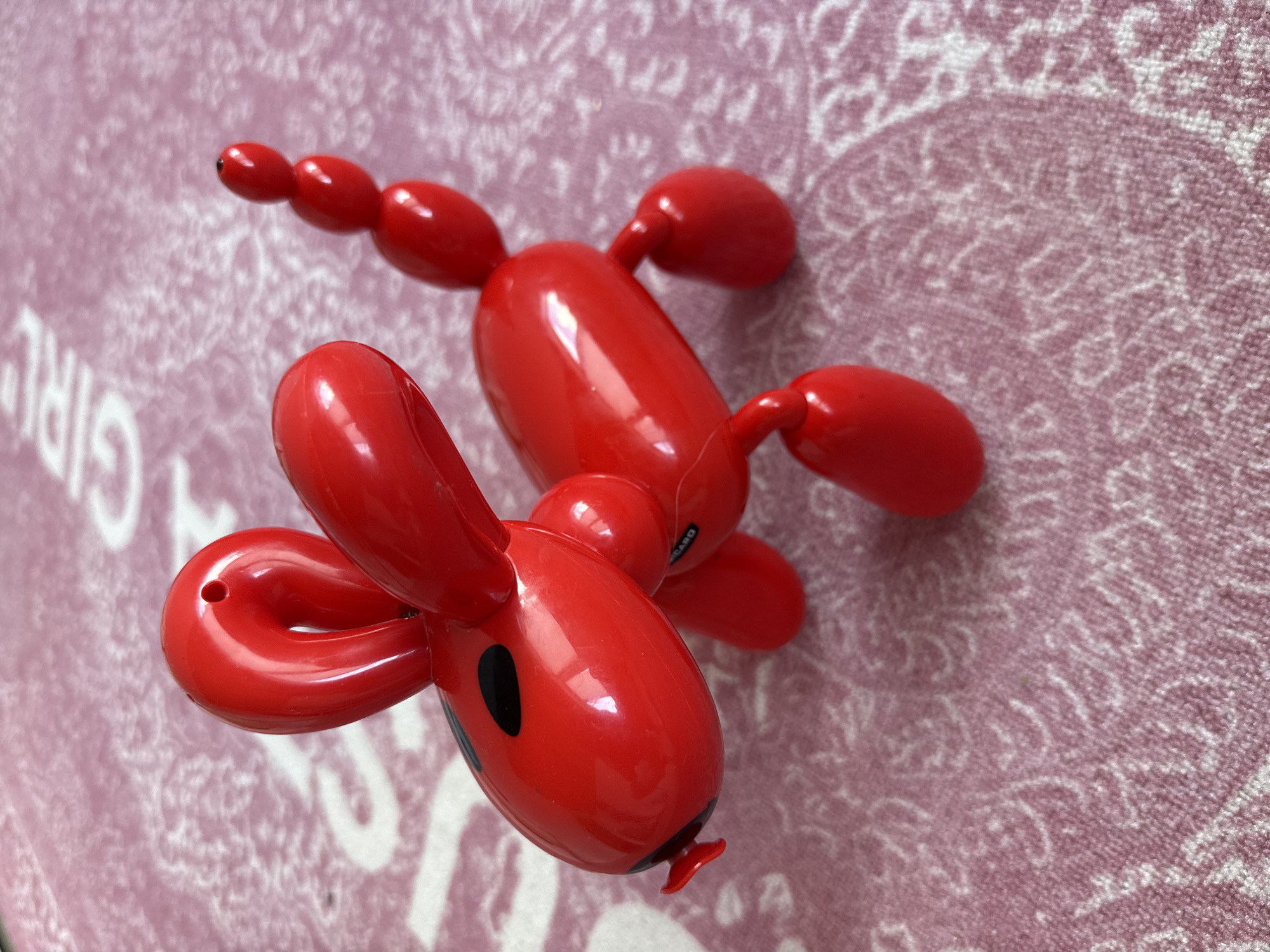 A close-up photograph depicts a red, shiny, plastic toy resembling a balloon animal dog. The toy is positioned diagonally on a surface with a pink and white patterned covering. The head of the dog has two looped ears and a black oval marking, possibly representing an eye. The body of the toy is elongated with several bulbous segments. It has limbs on both sides. The background is blurred. The exact location cannot be determined from the image, and there is no indication that the photo was taken in Votkinsk, Russia.