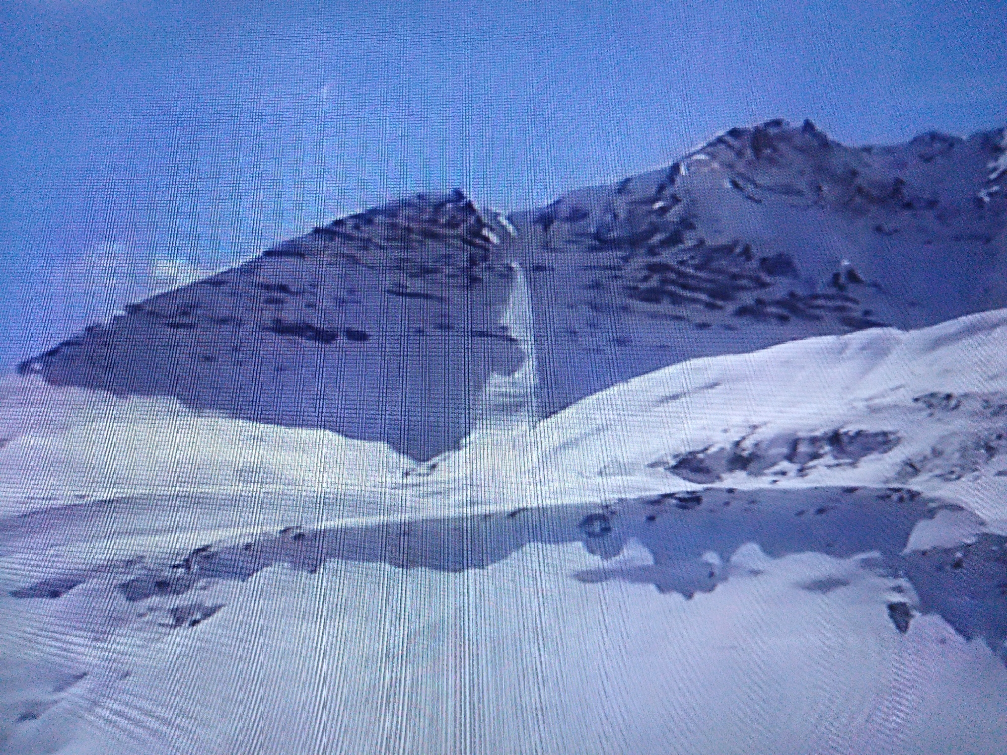The image depicts a panoramic view of a snow-covered mountain range under a clear blue sky. The mountains are rugged and appear to be a remote, natural landscape. There is no indication of any urban setting, buildings, or people. The scene is characterized by vast expanses of snow and rock formations. The image quality is low, appearing pixelated, which may be due to digital compression or capture limitations. There is no information in the image that would suggest a location in the city of Dushanbe, Tajikistan, or any other specific urban area. The environment is purely natural and mountainous.