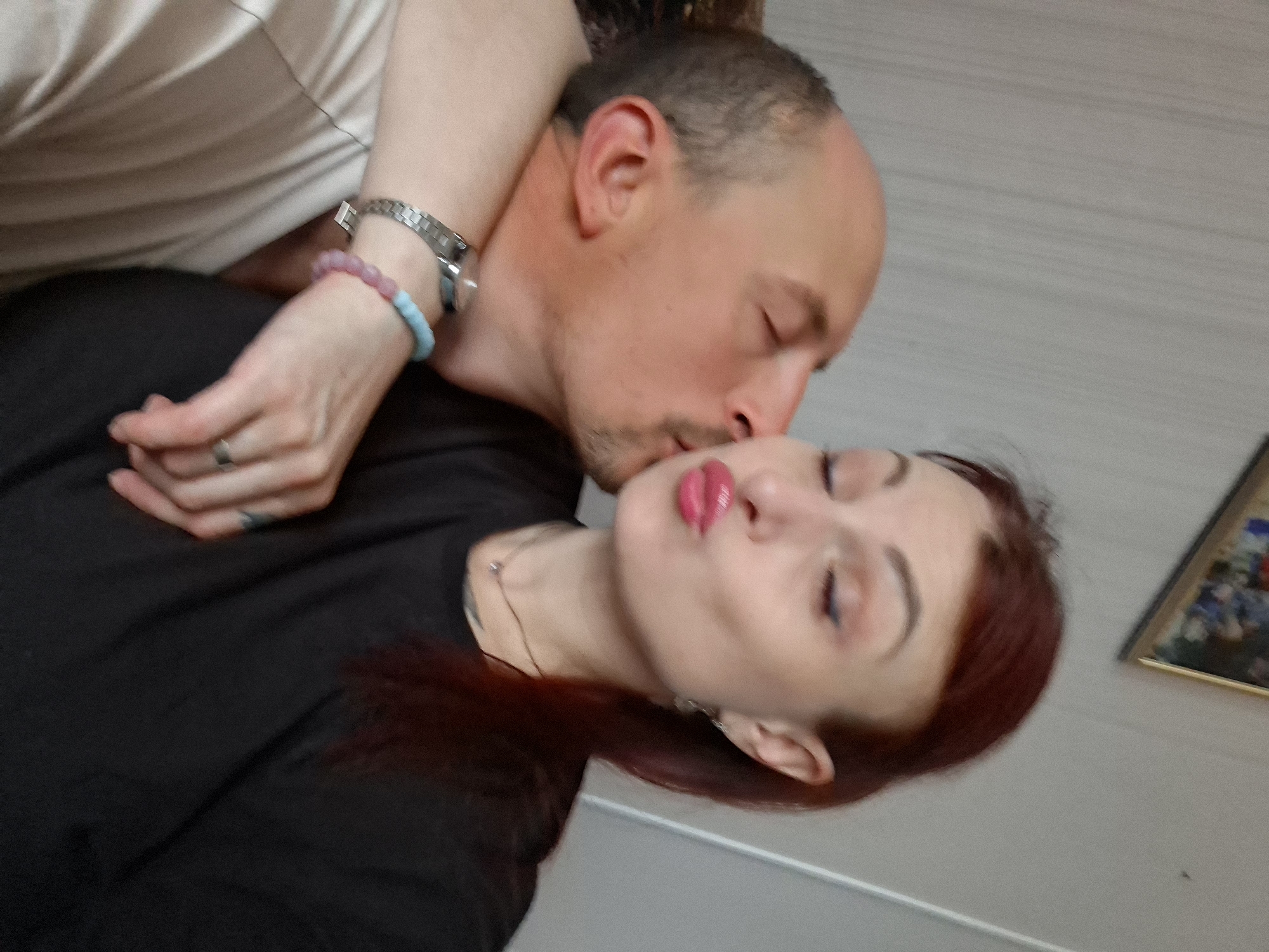 A close-up, indoor image depicts a man on the left kissing a woman on her puckered lips, both with their eyes closed. The man has a receding hairline or very short hair, dark facial stubble, and wears a light-colored t-shirt. A silver-toned watch with a metallic link strap is visible on his left wrist. The woman, positioned on the right, has reddish-brown hair, wears a black t-shirt, and has visible makeup including blue winged eyeliner and pink lipstick. She wears a thin silver-toned necklace with a small pendant, a silver-toned earring on her left ear, and on her right wrist, a beaded bracelet with light blue and pink stones alongside a plain silver-toned ring on her right ring finger. Her left arm is visible around the man's neck/shoulder. The background consists of a light-colored wall with faint vertical striations. A framed picture is partially visible on the wall in the upper right quadrant. The image provides no visual evidence to confirm the specified location of Забалоцкі сельскі Савет, Belarus.