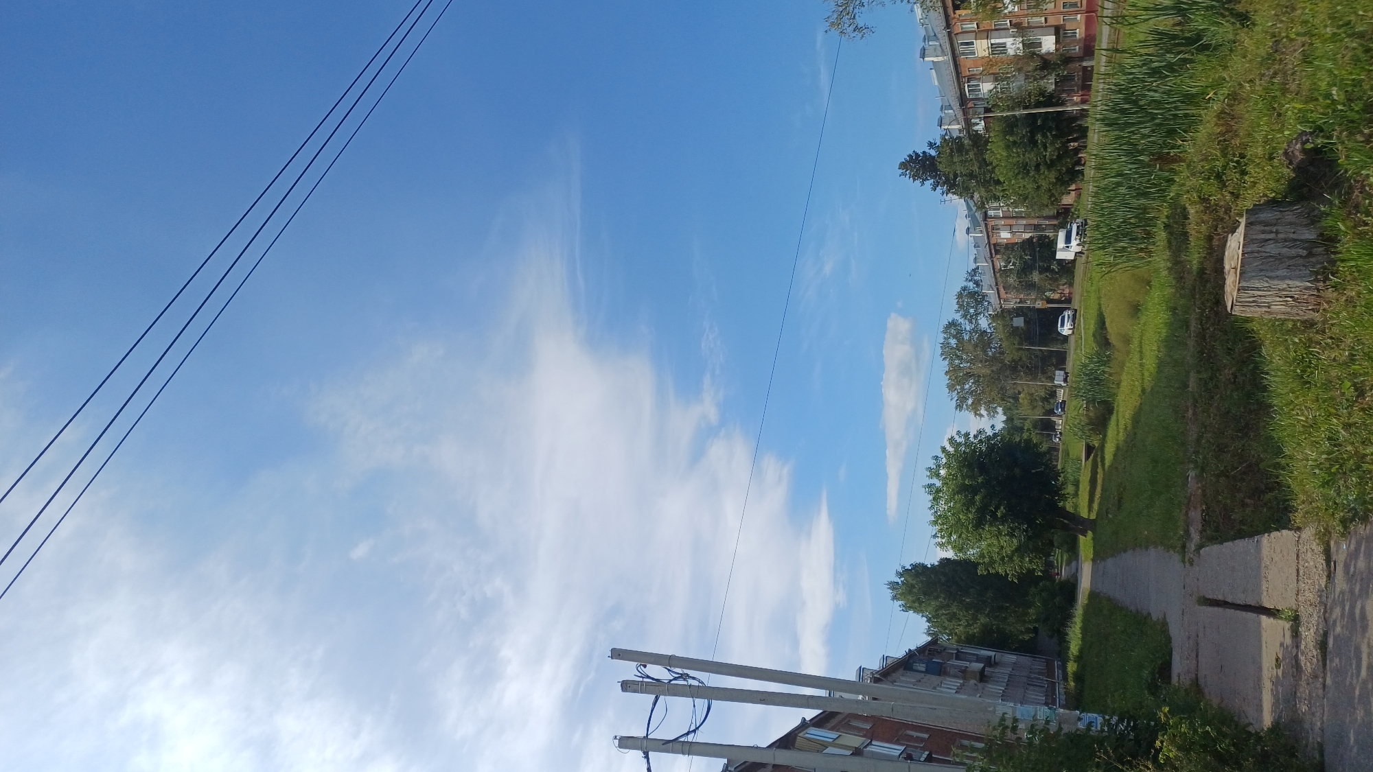 The image depicts a street scene in Izhevsk, Russia on a clear day with a blue sky and scattered white clouds. The perspective is from a sidewalk, looking down a tree-lined street. On the left, there is a multi-story brick apartment building with several windows. Adjacent to the sidewalk are concrete utility poles with electrical wires. The sidewalk itself is made of concrete slabs and shows signs of wear. To the right of the sidewalk is a grassy area with a tree and a large tree stump in the foreground. Further down the street, there are more apartment buildings and trees. Several vehicles, including white vans and cars, are visible on the road. Tall reeds or grasses grow along the side of the road. The overall impression is of a residential urban environment.