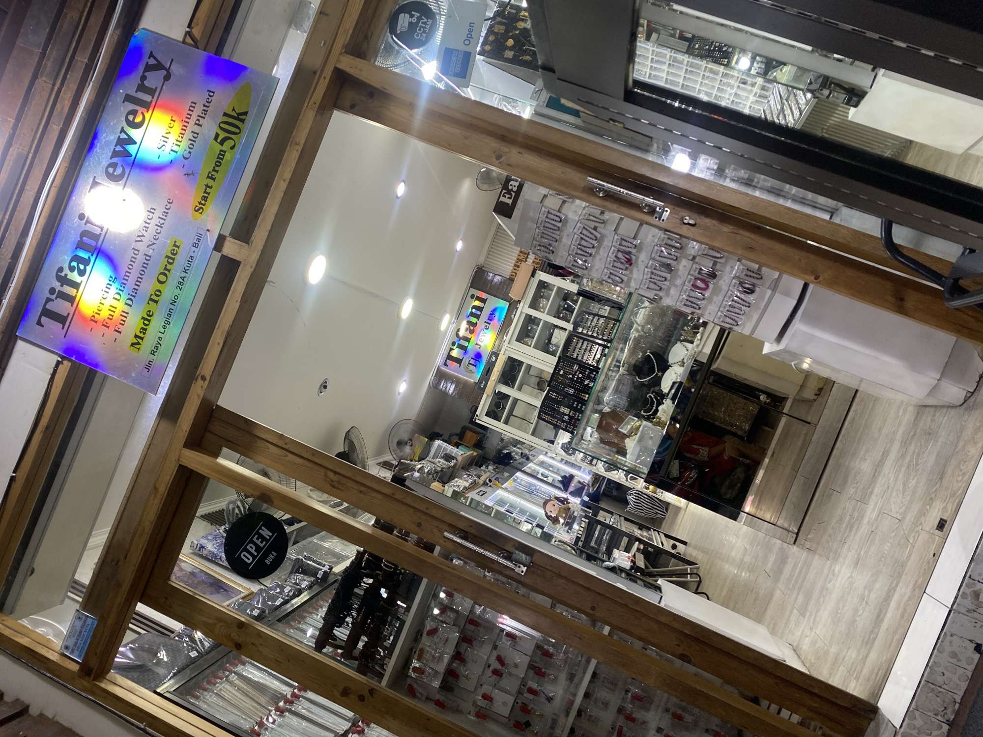 This image captures the interior of a jewelry store named "Tifani Jewelry" or "Tifani TJ Jewelry," viewed from just outside the entrance at an angled perspective. The store appears brightly lit and ready for customers.
The main sign prominently displays "Tifani Jewelry" and lists various offerings including "Piercing," "Full Diamond Watch," and "Full Diamond Necklace." The materials used are specified as "Silver," "Titanium," and "Gold Plated." Products are "Made To Order" and "Start From 50k." The full address, "Jln. Raya Legian No. 28A Kuta - Bali," is also visible on this sign, confirming the location context.
Inside the store, various jewelry items are displayed in glass cases and on shelves. There are numerous earrings, necklaces, and what appear to be watches or bracelets arranged in an organized manner. The shop features wooden framework around its entrance and display areas. Two ceiling fans and several recessed lights illuminate the space.
Other visible signs indicate "OPEN" (with "BUKA" in Indonesian on one), an "AMERICAN EXPRESS" logo, and a "CCTV 24 JAM" sign, suggesting security monitoring. The floor has a light wooden or laminate finish. The shop appears unoccupied at the time the photo was taken.