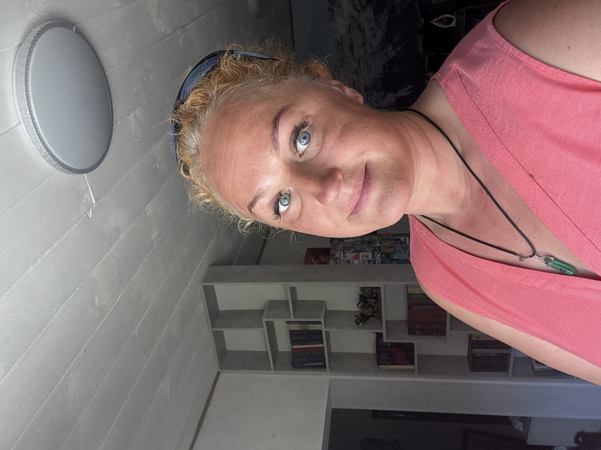 A woman with blonde, curly hair and blue eyes is shown in a close-up, head-and-shoulders shot. She is wearing a pink sleeveless top and a black necklace with a green pendant. Her eyes are looking towards the camera with a slight smile. The background appears to be an interior space with light-colored, horizontally lined walls. A circular light fixture is visible on the upper left. A bookshelf with several shelves filled with books and decorative items is visible in the background on the right. The lighting suggests it could be daytime. The image is oriented sideways.