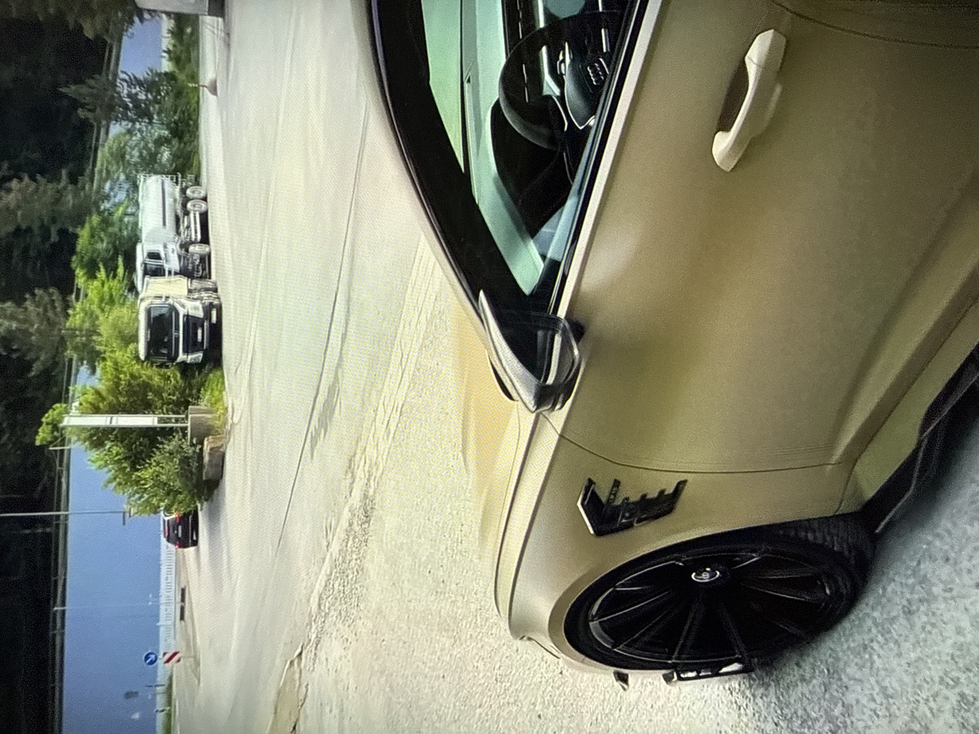 The scene is set outdoors in Varna, Bulgaria, depicting an angled view of a road and its surroundings. In the foreground, on the right side, a modified Audi RS6 station wagon is prominently displayed. The vehicle is finished in a matte gold or bronze color, equipped with multi-spoke black alloy wheels, and features an "ABT" branded vent on the front fender. Carbon fiber side skirts are visible along the lower body line. The driver's side mirror has a dark housing, and through the window, an Audi-branded steering wheel is discernible in the dark interior.
The midground features an asphalt road surface with visible texture and faint markings. A blue circular sign indicating a mandatory right turn and a red and white striped triangular warning sign are present on the left side of the road. Further into the midground, a dark-colored SUV or sedan is partially visible, appearing to approach the intersection. On the right side of the road, two large commercial trucks are parked: a white Volvo semi-truck cab and, behind it, a white tanker truck.
The background consists of dense green foliage and trees, behind which a long, light blue wall or structure is visible. No individuals are present in the image. The entire image exhibits a digitized, pixelated quality.
