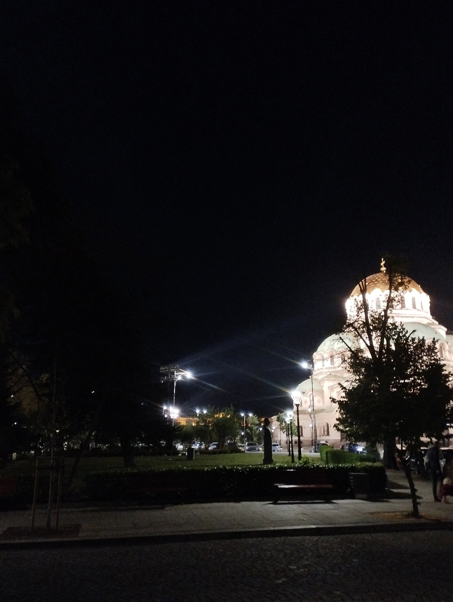 The image presents a nighttime view of a large, illuminated building, likely an Eastern Orthodox cathedral or church, set against a dark sky. The dominant architectural feature is the building's distinct domes; a prominent central dome glows with a golden-bronze hue, topped by a cross, while another, partially visible dome, appears to have a green or teal finish. The cathedral's white facade is brightly lit, revealing architectural details such as arched windows.
In the foreground and midground, a park or garden area is visible, characterized by manicured grass, hedges, and trees, some of which are silhouetted against the lighter background of the building. Several streetlights cast bright illumination, with some producing noticeable lens flares. A dark, paved surface, resembling a sidewalk or street, spans the bottom of the frame. Within the park, a wooden bench is positioned, and a dark statue or bust on a pedestal is discernable on the right side.
A few indistinct human figures, including what appears to be an adult and a child, are visible on the far right, suggesting people are strolling through the public space. The scene emanates a calm and peaceful ambiance, typical of an urban setting at night. There is no visible text in the image.