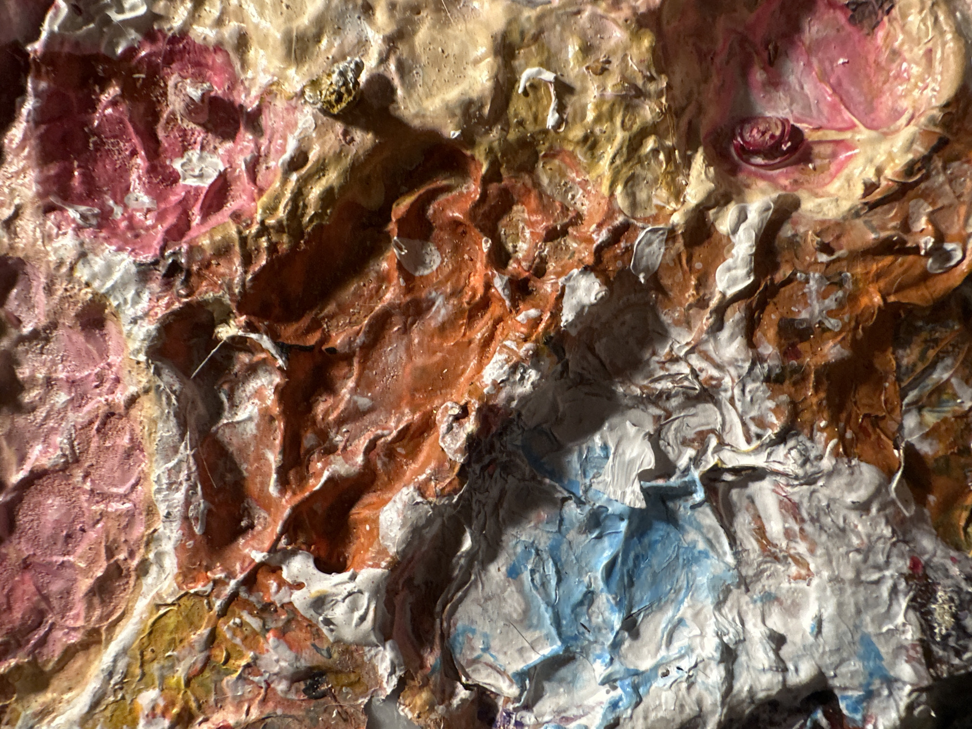 The image displays a highly textured close-up of an abstract painting. The paint is applied thickly, creating a sculptural, impasto effect with visible ridges, swirls, and peaks. The colors are a vibrant mix of warm tones like orange, pink, and gold, contrasted with cool blues and whites, and areas of deep shadow. The texture suggests a sense of depth and dynamism, with light reflecting off the raised surfaces. There are no discernible people, objects, or specific environmental features visible in this abstract composition. The image is a detail shot of the artwork itself, not a depiction of a location in Yekaterinburg, Russia.