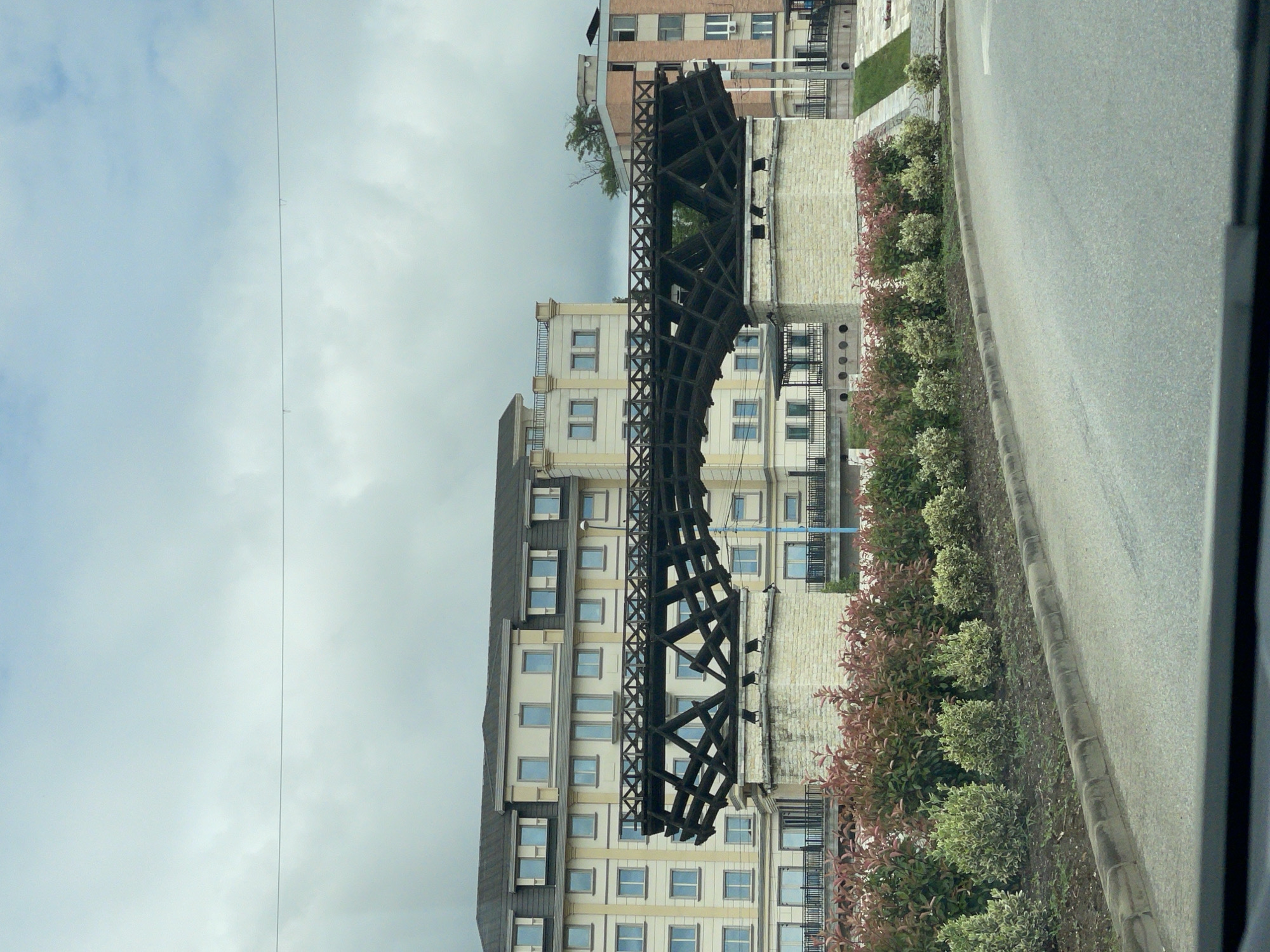 The image displays an urban landscape featuring a large, dark, intricately latticed metal structure that curves through the middle ground. This structure, which resembles a segment of Trajan's Bridge, stands prominently in front of several light-colored, multi-story buildings with numerous rectangular windows and some balconies.
In the foreground, a paved asphalt road runs along the right side of the scene, bordered by a concrete curb. Adjacent to the curb is a landscaped strip of ground planted with rows of green and reddish-leafed bushes. The upper portion of the image is dominated by a cloudy, light-gray sky, traversed by a thin, barely visible wire or cable.
The scene depicts a daytime setting under an overcast sky. There are no people visible, and no specific activities are taking place. No text is discernible within the frame. The architecture and the distinctive metal structure are consistent with an urban environment, specifically suggesting Drobeta-Turnu Severin, Romania, given the historical significance of Trajan's Bridge in that region.