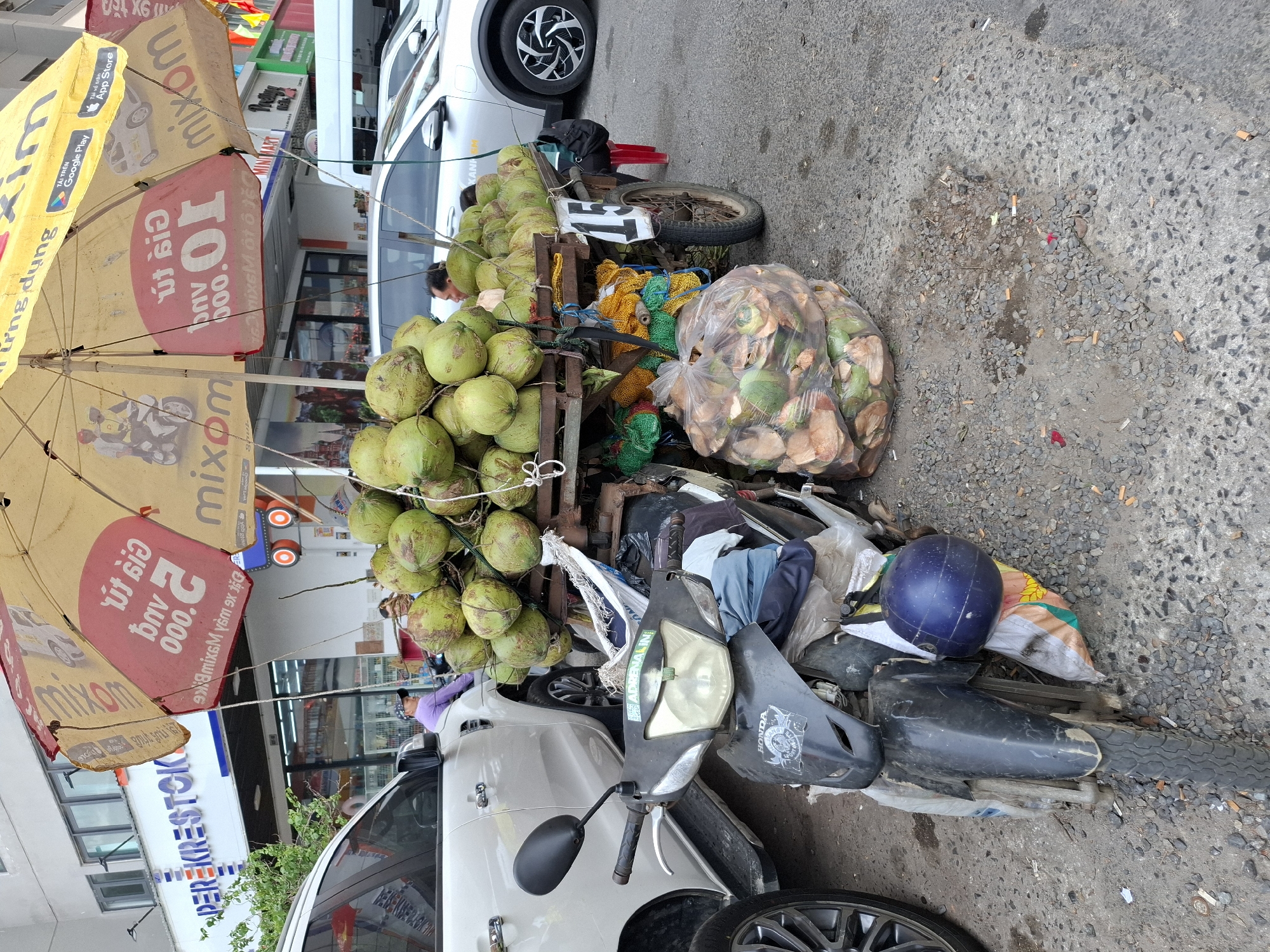 In Nha Trang, Vietnam, a street vendor operates a mobile coconut stand from a motorbike-drawn cart. The cart is laden with a large quantity of green coconuts, stacked high and secured with ropes. A large umbrella shades the coconuts, displaying promotional text and logos, including "mixom" and app store icons. In the foreground, a black motorbike is parked, its front end partially covered with various bags and clothing items, with a blue helmet resting on top. Behind the cart, several cars are visible, including a white SUV and a silver sedan. The scene is set on a paved street, with a building displaying signage for "PEREKRESTOK" in the background. The ground is littered with gravel and cigarette butts. A man is partially visible behind the coconuts, presumably the vendor.