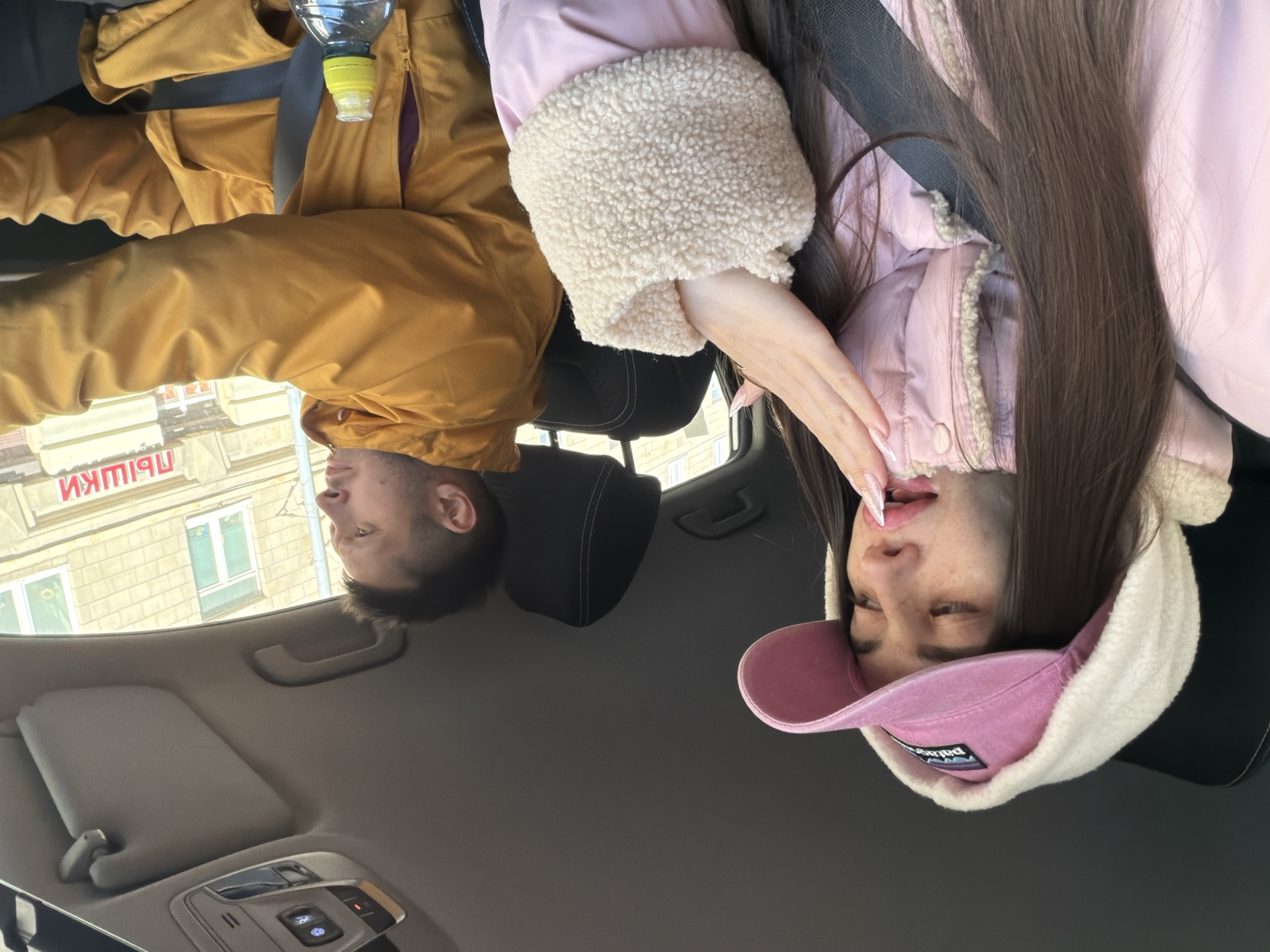 A photo taken inside a car, shows two people wearing jackets. A man in a yellow jacket on the left and a woman wearing a pink jacket with a white fur hood and pink baseball cap on the right. In the background, a building with text in Russian which when translated to English reads "Torture". Likely taken in Saint Petersburg, Russia.