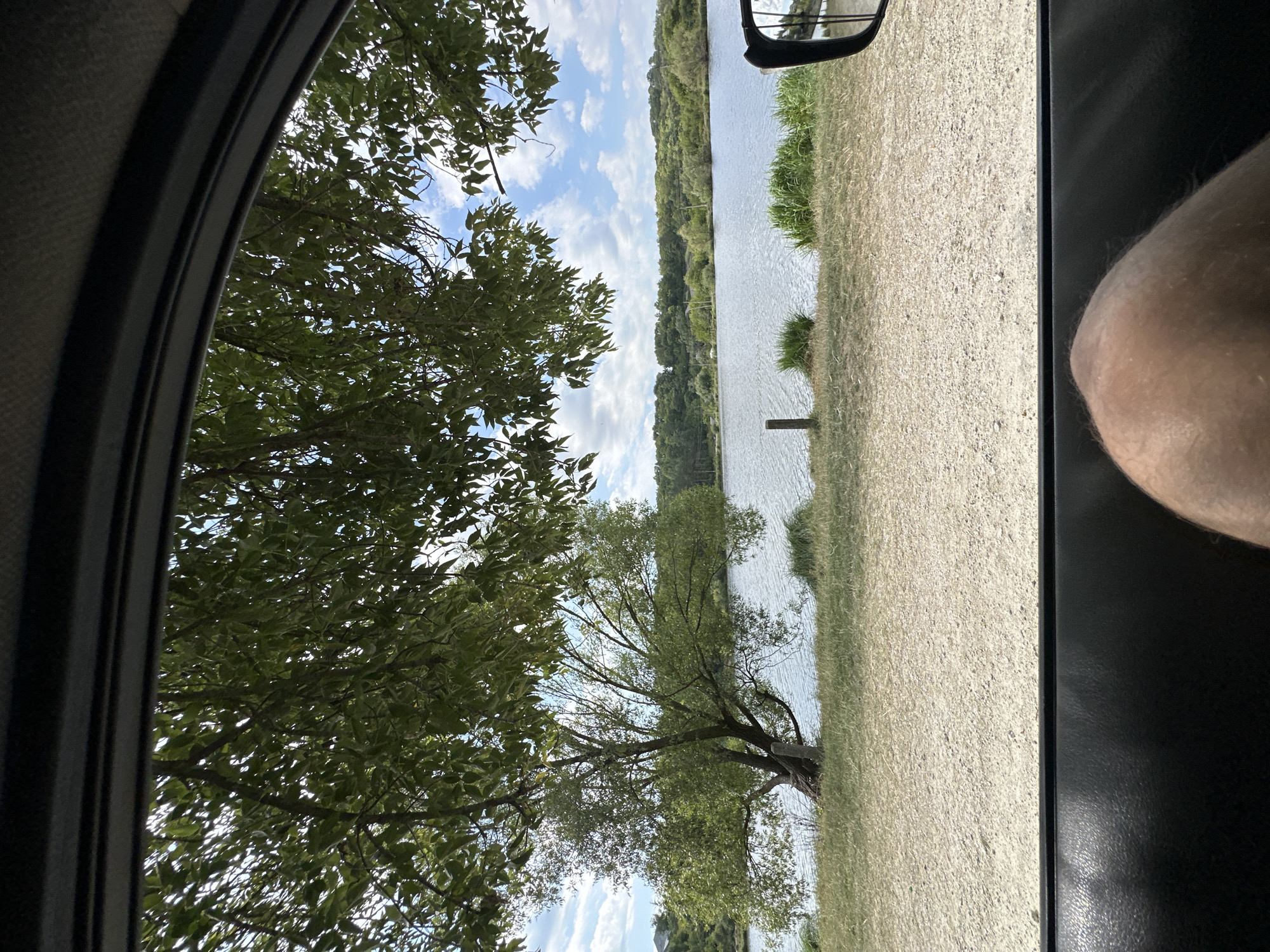 The image shows a view from inside a vehicle, looking out towards a body of water. The foreground is dominated by the interior of the car, including part of a dark leather door panel and a person's leg. Through the window, a tree with green foliage is visible, casting shadows. Beyond the tree, a calm lake or river stretches across the frame, with a tree-lined shore in the distance. The sky is partly cloudy with blue patches. The environment appears to be a natural outdoor setting, possibly a park or waterfront area. The location in the city of Makiv, Ukraine is not explicitly discernible from the visual information alone.