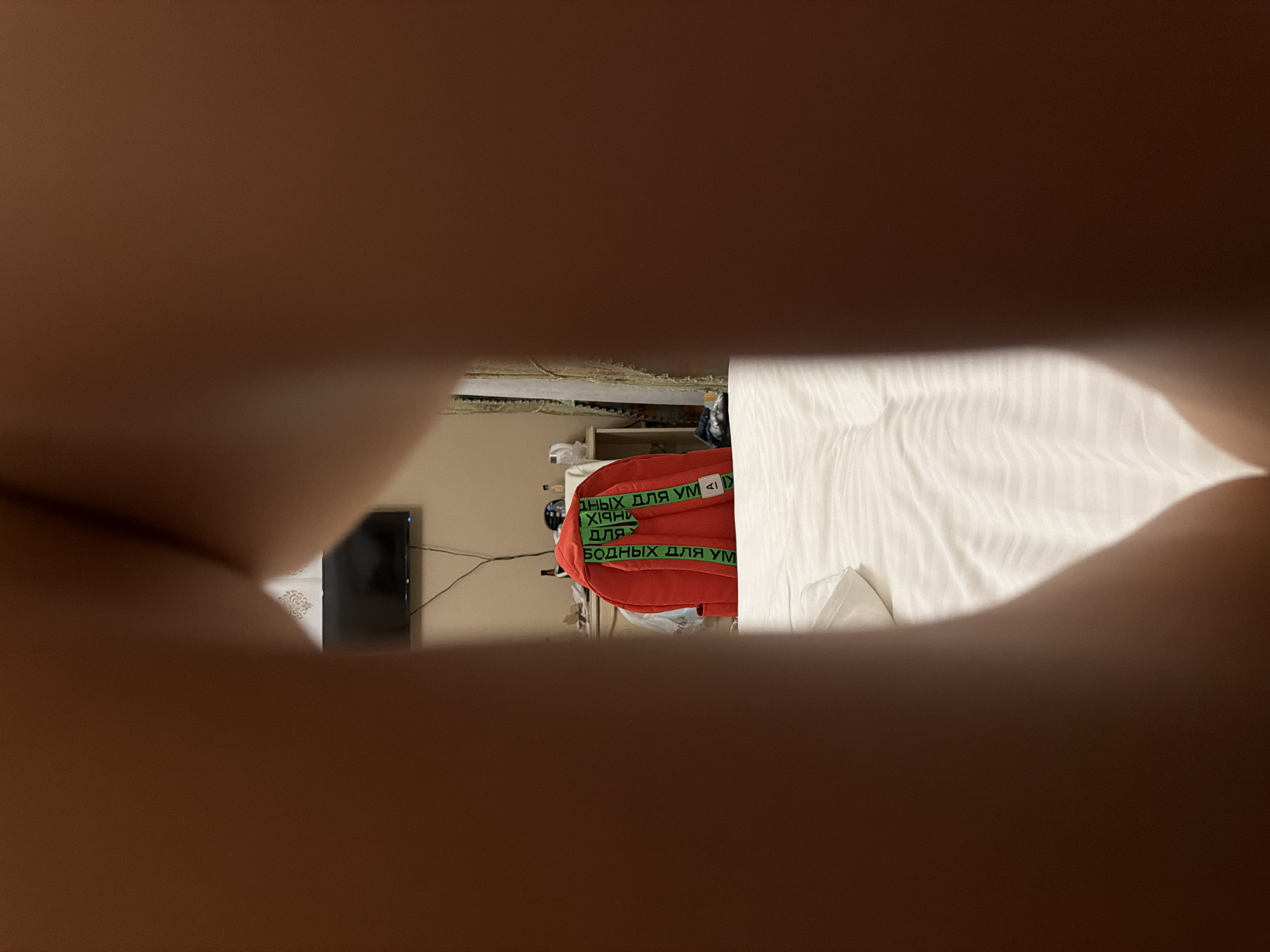 The image appears to be taken from a low angle, looking through a gap formed by what seem to be legs. The foreground is dominated by blurred, brown-toned skin.
In the background, a red backpack with green straps is visible. The straps have text printed on them, which appears to be in Cyrillic script. The text "НЕ ПРЫГАЙ ДЛЯ VM" (Do not jump for VM) and possibly other similar phrases are discernible on the straps.
Behind the backpack, a dresser or table is present, with various items on it, including what looks like a bottle and a white object, possibly a pitcher or kettle. Further back, a television screen is mounted on the wall. Curtains are visible on the right side of the background. The setting appears to be an indoor space, possibly a bedroom, with a neatly made bed covered in white linens in the mid-ground.
The location is stated to be Zelenogradsk, Russia. However, there are no specific visual cues in the image that definitively place it in Zelenogradsk. The scene is consistent with a hotel room or a domestic bedroom.