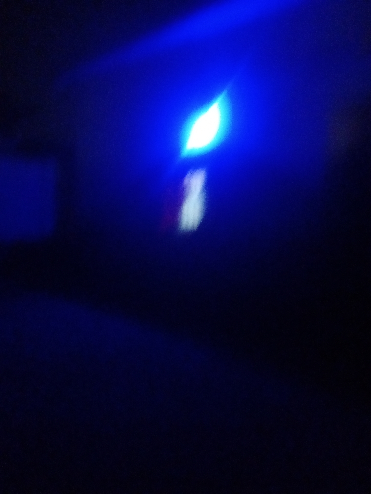 The image shows a blurred, dark environment illuminated by a bright blue light source. The light appears to be emanating from the top left of the frame, casting a beam that illuminates a small, white, leaf-like shape in the center of the image. There are no discernible people, specific objects, or actions visible. The setting is too dark and indistinct to determine if it is indoors or outdoors, or to provide any specific details about the location in Sabon Gari, Nassarawa, Nigeria. The overall impression is one of abstract light and shadow.