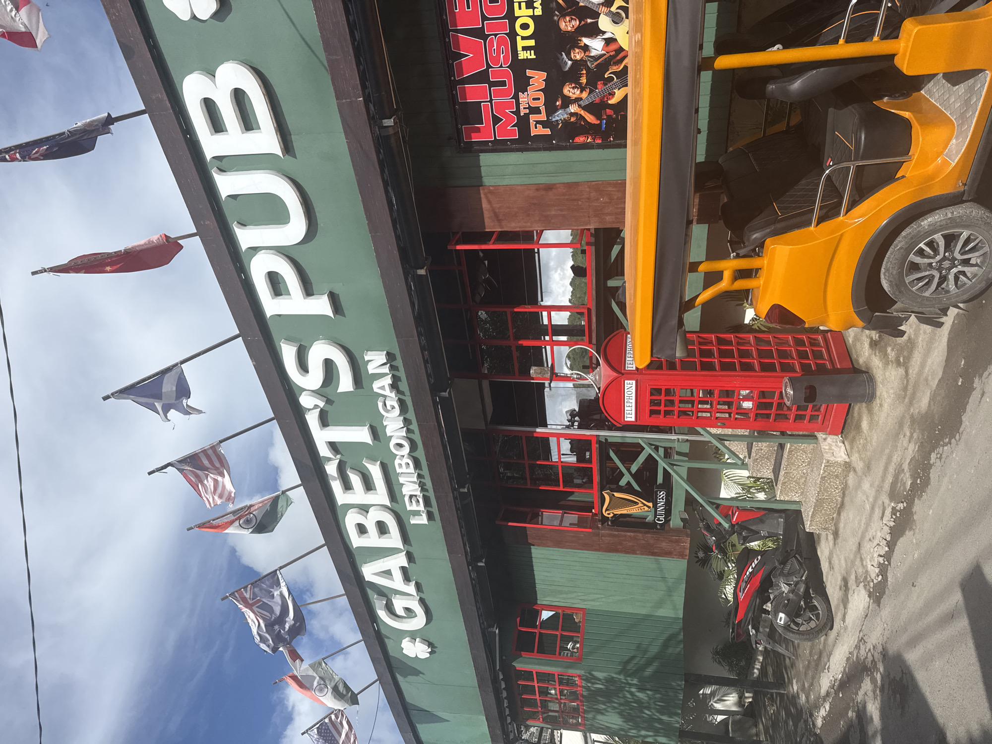 The image captures the exterior of Gabet's Pub Lembongan, located on Nusa Penida, Indonesia. The pub features a green facade with red trim on windows and doors, as well as a red British telephone booth. A black and red motorcycle is parked near the entrance. Flags of various nations are displayed above the pub. A yellow vehicle with black seating is partially visible on the right. A poster advertising "Live Music The Flow" is placed above the pub entrance. The sky is visible with scattered clouds.