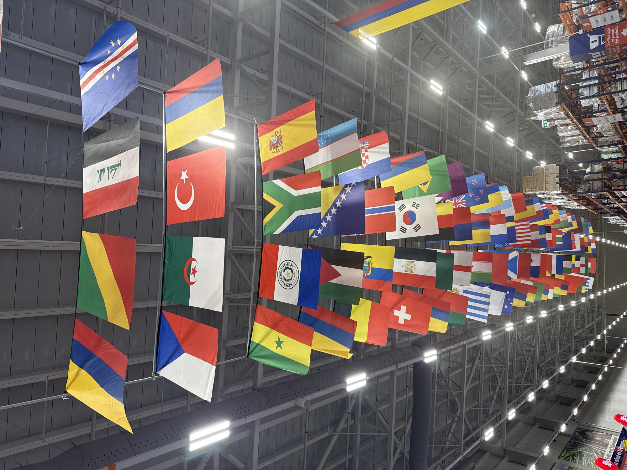 The image shows an interior view of a large warehouse-style building, possibly a retail store or distribution center, with a high ceiling supported by exposed metal beams and trusses. Fluorescent lights are mounted on the ceiling, illuminating the space. The primary feature of the image is a large display of numerous national flags suspended from the ceiling by wires, creating a colorful canopy.
The flags are of various sizes and are hung in rows, overlapping and creating a dense visual effect. Some of the visible flags include those of Colombia, Congo, Czech Republic, Algeria, Iraq, Cape Verde, Turkey, Paraguay, Senegal, South Africa, Spain, Bosnia and Herzegovina, South Korea, Brazil, and others. The flags are in different orientations, some hanging straight down, while others are angled.
In the background, industrial shelving units are visible, filled with boxes and pallets of merchandise, indicating a commercial or logistical purpose for the building. The overall environment is utilitarian and vast.
The city of Mosquera, Colombia is not directly depicted or indicated within the image itself. The context of Mosquera, Colombia is provided as the location of this scene, implying that the warehouse is situated there.