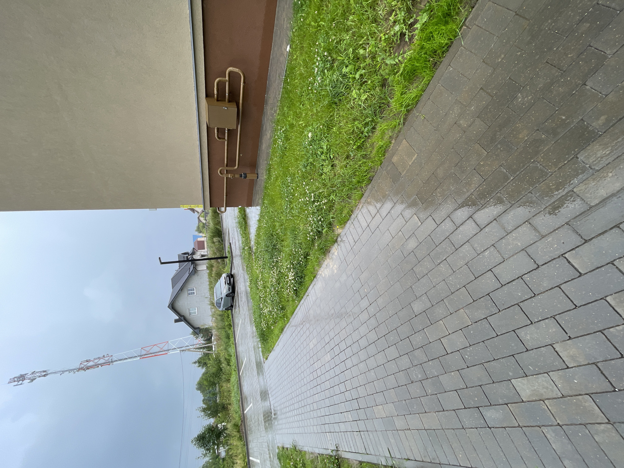 The image shows a wet, paved walkway in Zelenogradsk, Russia, leading towards a parking lot and a residential building. A grey car is parked in the lot next to a grassy area with small white flowers. In the background, a tall, red and white telecommunications tower stands against a cloudy sky, with a house and trees visible between it and the residential building. The residential building features a light beige facade with brown trim near the ground, where brown pipes and a small utility box are visible. The paved walkway is made of dark grey rectangular stones. The scene appears to have been recently rained on, as evidenced by the wet pavement and a generally overcast sky.