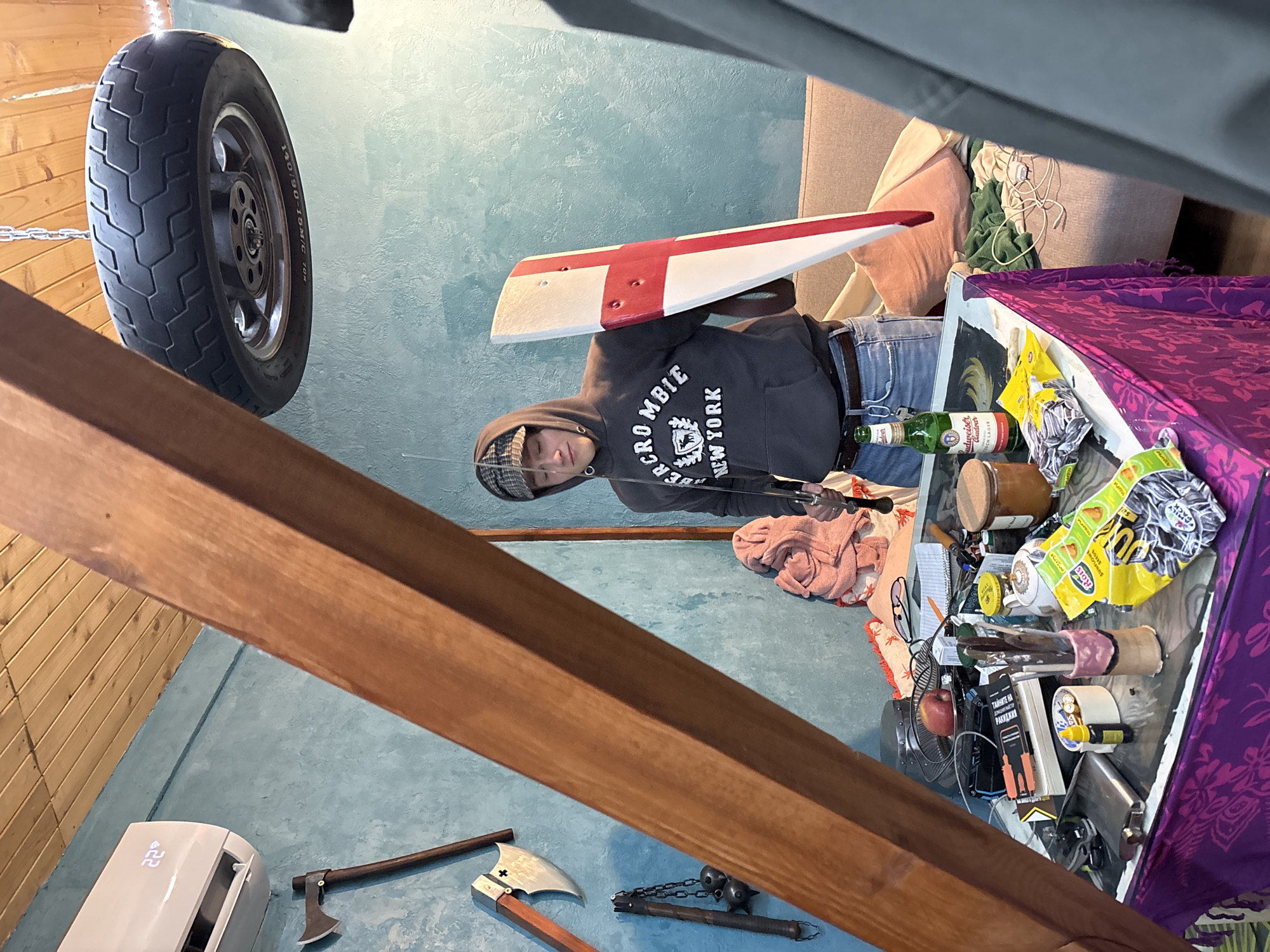 The image depicts an indoor scene within a room with a light blue wall. A person wearing a grey hooded sweatshirt, jeans, and a plaid cap is holding a white object with a red cross design. The room is cluttered with various objects, including a tire hanging from the ceiling, a wooden beam, an air conditioning unit, axes, a flail, a table covered in a purple patterned cloth, a couch, a bottle of Budweiser beer, food packaging ("ROLS"), and other miscellaneous items. The book says "РАКИДКИЯ ТАЙНИТЕ НА".