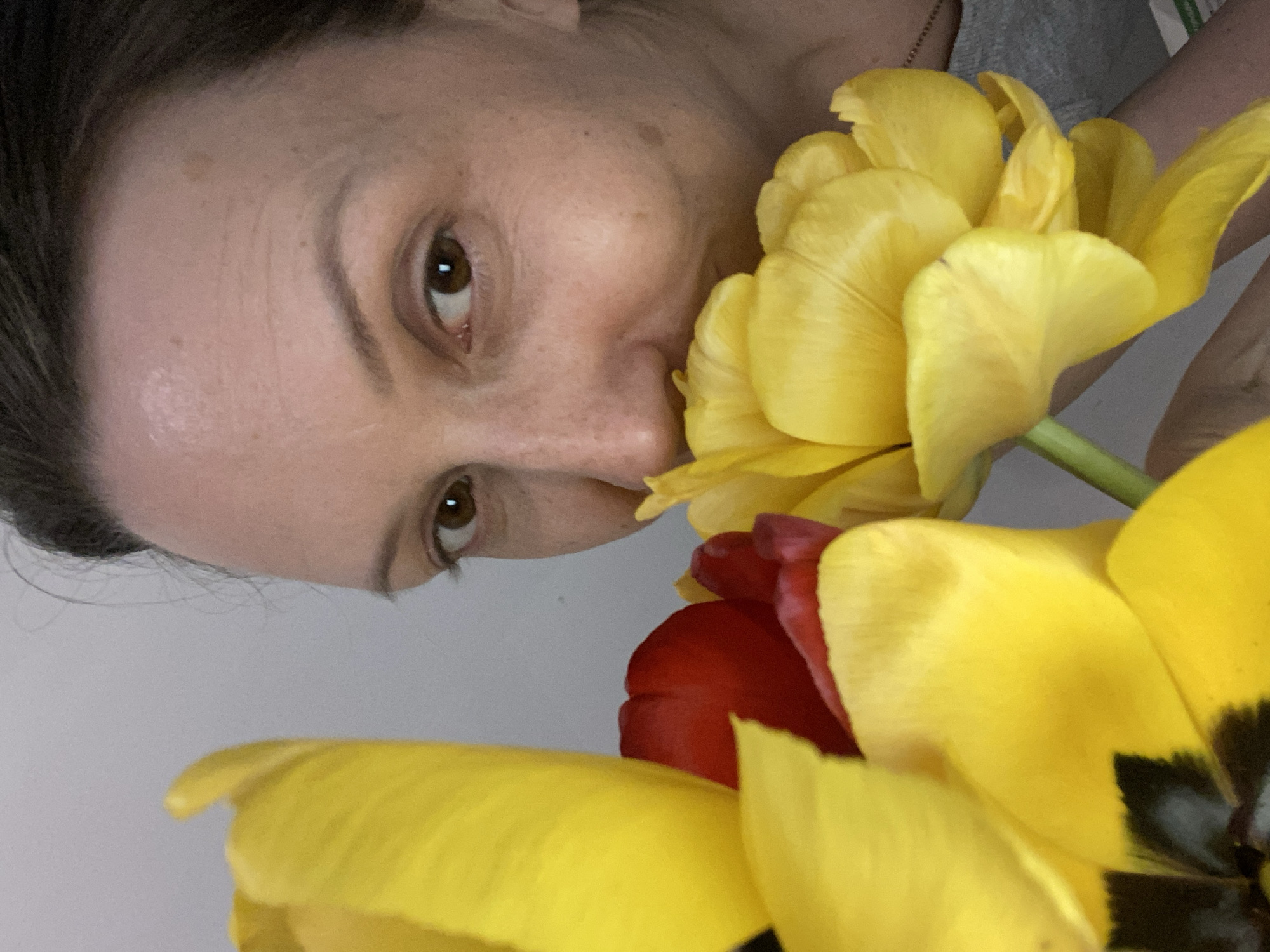 A woman with brown hair and eyes is holding yellow and red tulips close to her face. The background is a plain white wall. It is not possible to determine if the photo was taken in the city of Chaikovsky, Russia, based on the visual information provided.