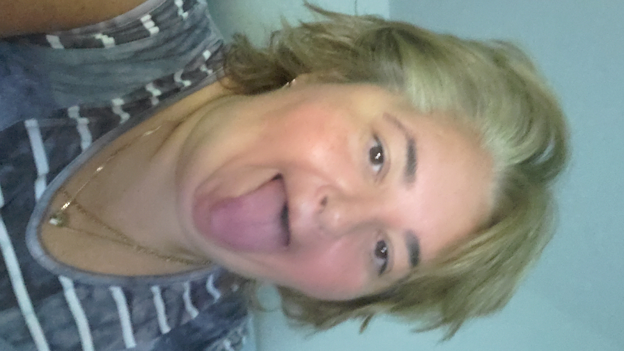 A woman sticks her tongue out at the camera in a selfie. She has blonde hair, and is wearing a striped shirt and a necklace. She is indoors, with a plain blue wall in the background. The image was taken at the Baden-Airpark, an airport located in Rheinmünster, near Baden-Baden, which is in the state of Baden-Württemberg, Germany.