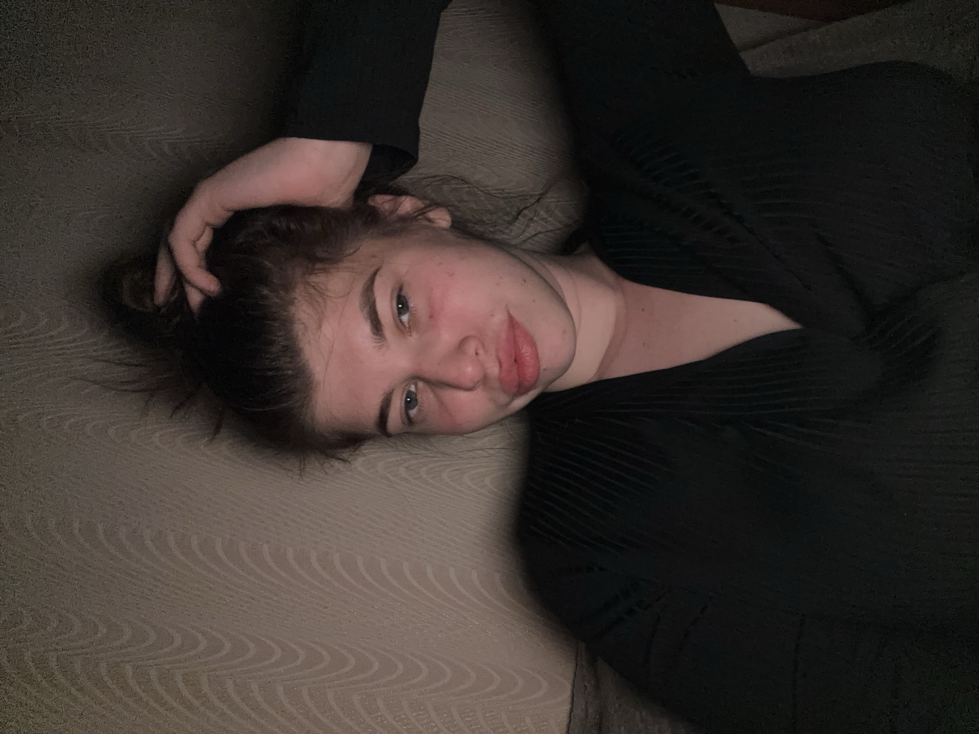 A young woman is lying on her back on what appears to be a patterned couch or bed cover. She is wearing a dark top with a ribbed or textured pattern. Her right hand is placed on top of her head, her hair is gathered up. Her face is slightly flushed, and she is looking directly at the camera. The lighting is dim. No indication of location.