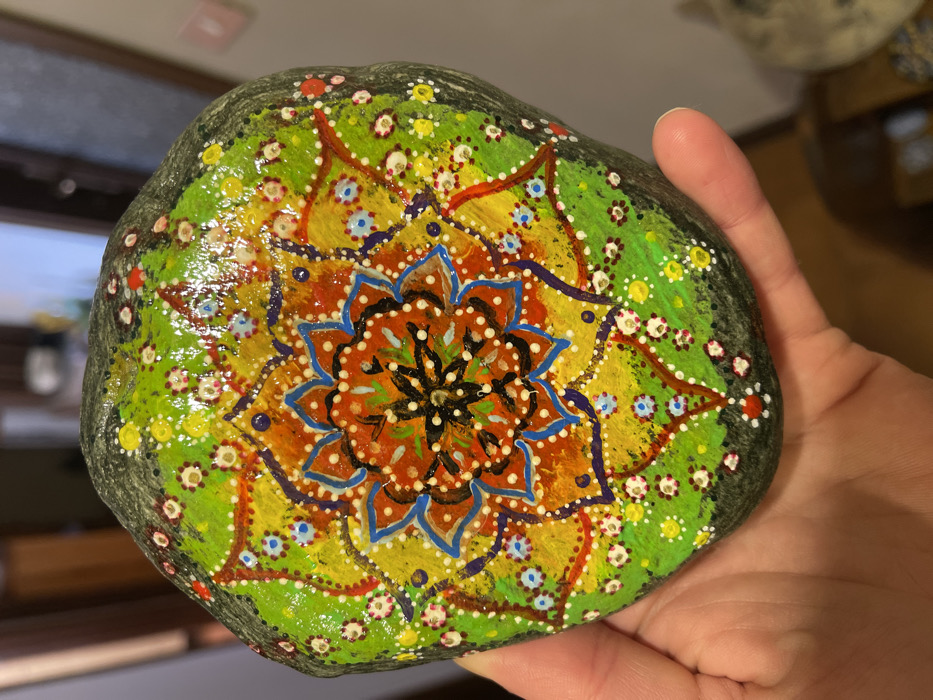 A hand holds a brightly painted, smooth, oval-shaped rock indoors. The rock is adorned with an elaborate, mandala-style design that covers its entire top surface. At the center, there's an intricate, multi-layered motif in shades of orange and brown, resembling a stylized flower or star, accented with blue outlines and white dots. This central pattern is surrounded by a ring of vibrant yellow, which features smaller, dotted star shapes and thin purple lines. The outermost painted layer is a lively lime green, richly embellished with numerous small white, red, and yellow dots that create a textured, speckled effect. The painting has a glossy, sealed finish that reflects light, highlighting the intricate details. The hand, light-skinned with visible knuckles, supports the rock, which is substantial enough to nearly fill the palm. The background is softly blurred, showing indistinct shapes and colors characteristic of an indoor setting, suggesting artificial lighting. No text or specific location cues are visible within the image.