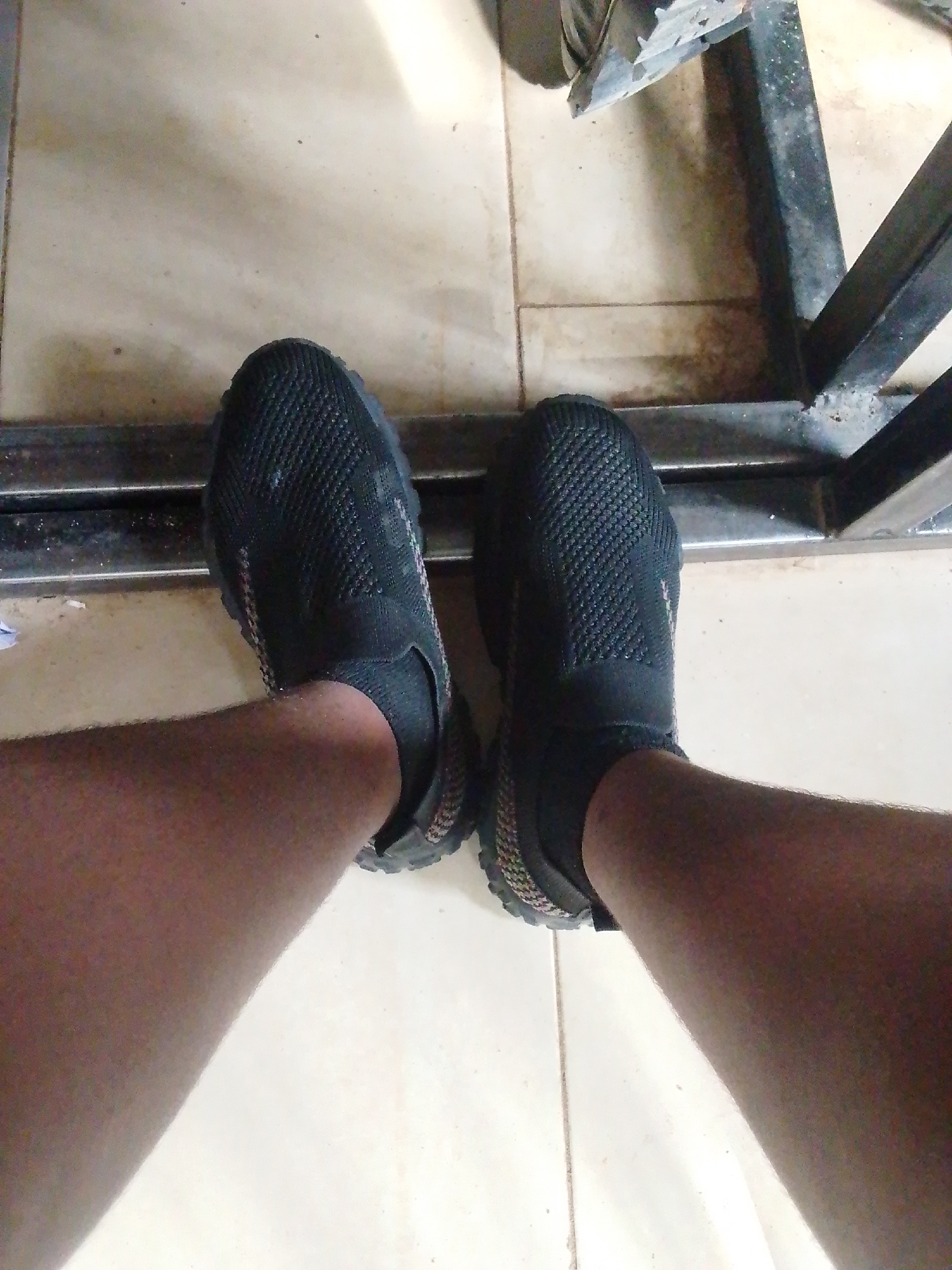 The image shows a person's legs and feet, clad in black slip-on shoes with a textured knit upper and a multi-colored sole. The person is standing on a tiled floor that appears to be in an outdoor or semi-outdoor setting, possibly a patio or walkway. A portion of a dark metal structure, likely furniture or part of a building, is visible in the background. The floor tiles are light-colored with visible grout lines. The overall lighting suggests it is daytime. This scene is located in Kuson, Nigeria.