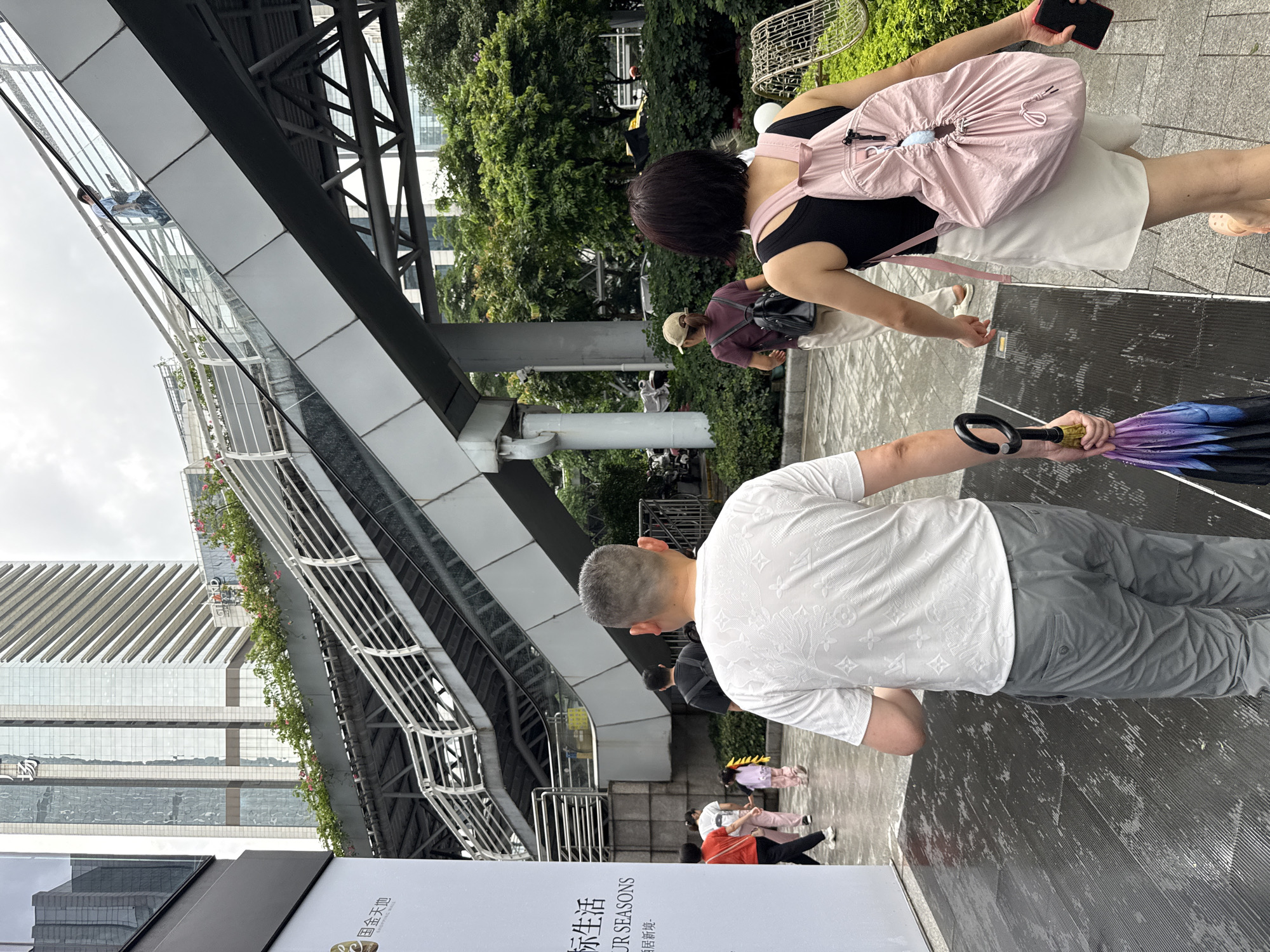 The image depicts an outdoor scene in Tianhe District, China, under an overcast sky. In the foreground, a man in a white patterned t-shirt and gray pants walks away from the camera, carrying a closed, multi-colored umbrella in his right hand. To his right, a woman in a black tank top and white skirt also walks away, wearing a light pink backpack and holding a red smartphone in her right hand.
Behind them, other pedestrians are visible. A woman in a maroon t-shirt and a tan baseball cap, wearing a black backpack, walks to the left. Further back, a woman in a white shirt and pink pants, and a young child in a pink outfit with yellow hair accessories, are walking towards the left. Another person in a dark t-shirt is visible walking away from the camera.
The environment includes a modern pedestrian overpass with metal railings and planted greenery. In the background, tall, glass-fronted buildings rise, characteristic of a city landscape. The ground in the foreground appears wet, suggesting recent rain or a damp surface, with a patterned texture. Lush green trees and bushes line the area to the right of the walkway. A white sign with Chinese characters and the text "OUR SEASONS" is partially visible on the left.