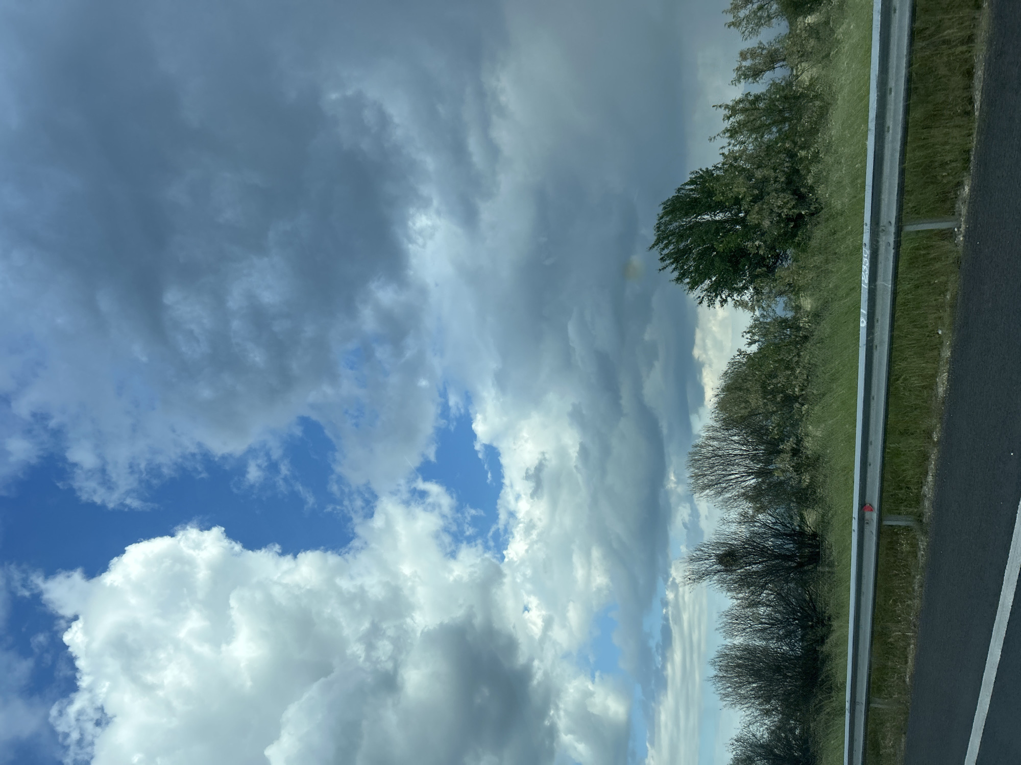 The image captures a roadside view under a dramatic sky in what could potentially be the vicinity of Malo Konare, Bulgaria, though the absence of specific landmarks prevents definitive confirmation. The composition is divided between the sky and the roadside environment. The sky dominates the frame, featuring a mix of dark, heavy storm clouds and patches of blue sky. Fluffy white cumulus clouds are visible in some areas, suggesting an unstable weather pattern.
The roadside features a metallic guardrail running parallel to a black asphalt road with a white line marking. Beyond the guardrail, there is a grassy area with a variety of trees and bushes of different sizes and densities. The scene lacks human presence.