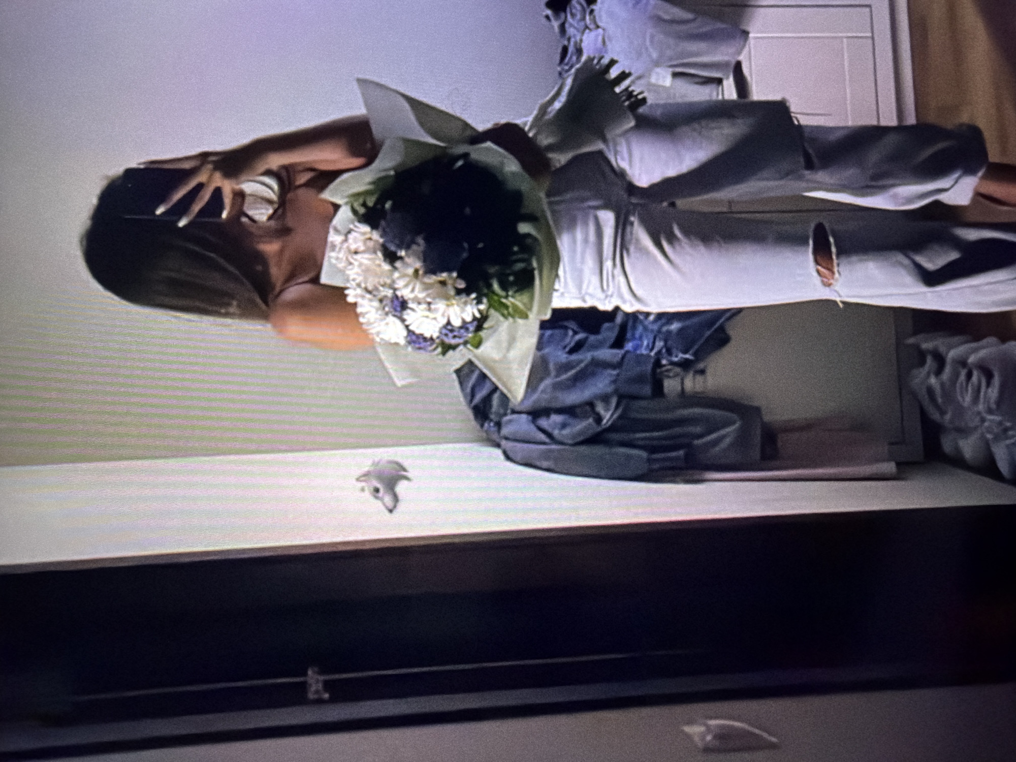 In what appears to be an indoor domestic setting, likely a bedroom or dressing area, a person is captured taking a mirror selfie. The individual, holding a bouquet of white and dark purple flowers wrapped in light green paper, is dressed in a light-colored spaghetti strap top and light-wash ripped jeans. The person's face is obscured by their phone, which they are using to take the photograph.
The environment shows a wall with vertical stripes, a dark-colored door or wardrobe on the left, and a dresser or cabinet on the right. Clothing, specifically denim jeans and other garments, are draped over the back of a chair or similar furniture. A small, decorative wolf figurine is visible on the wall to the left of the person. The overall impression is of a casual, personal moment captured within a lived-in space. The location is identified as Beșalma, Moldova.