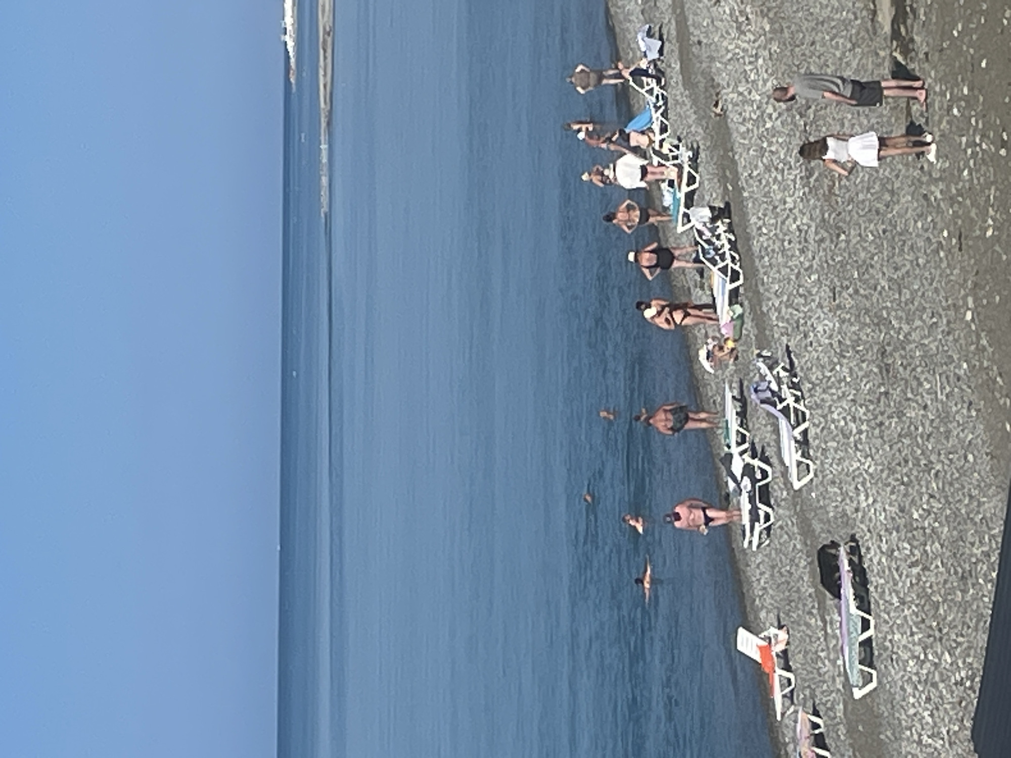 This is a high-angle, full-shot photograph of a pebble beach in Giachrypsh, Abkhazia. The scene captures a sunny day at the Black Sea, with a clear blue sky and calm, blue water. Several people are visible in the image, both on the beach and in the water.
On the beach, there are numerous empty and occupied lounge chairs scattered across the pebbles. Some individuals are standing, some are lying on the lounge chairs, and a few are walking. In the water, several people are swimming or wading.
In the foreground, two people are standing on the beach: a woman in a white dress and sneakers, and a man in a grey t-shirt and dark shorts. Behind them, a group of people are gathered near lounge chairs, some dressed in swimwear. Further down the beach, more individuals are seen relaxing on lounge chairs or standing.
The water itself shows several swimmers at various distances from the shore. The horizon line is clearly defined, with a faint line of land visible in the far distance. The overall impression is one of a typical summer day at a seaside resort.