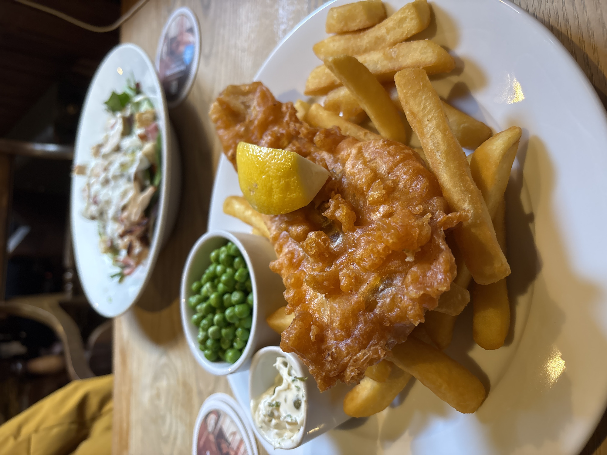 The image shows a plate of fish and chips. The fish is battered and fried, and a wedge of lemon sits on top. French fries are on the side of the fish. Additionally, there is a small bowl of peas and a small bowl of tartar sauce. To the left, there is also a small salad. The scene appears to be set on a wooden table, and the setting may be a pub or restaurant in the Vale of White Horse, United Kingdom.