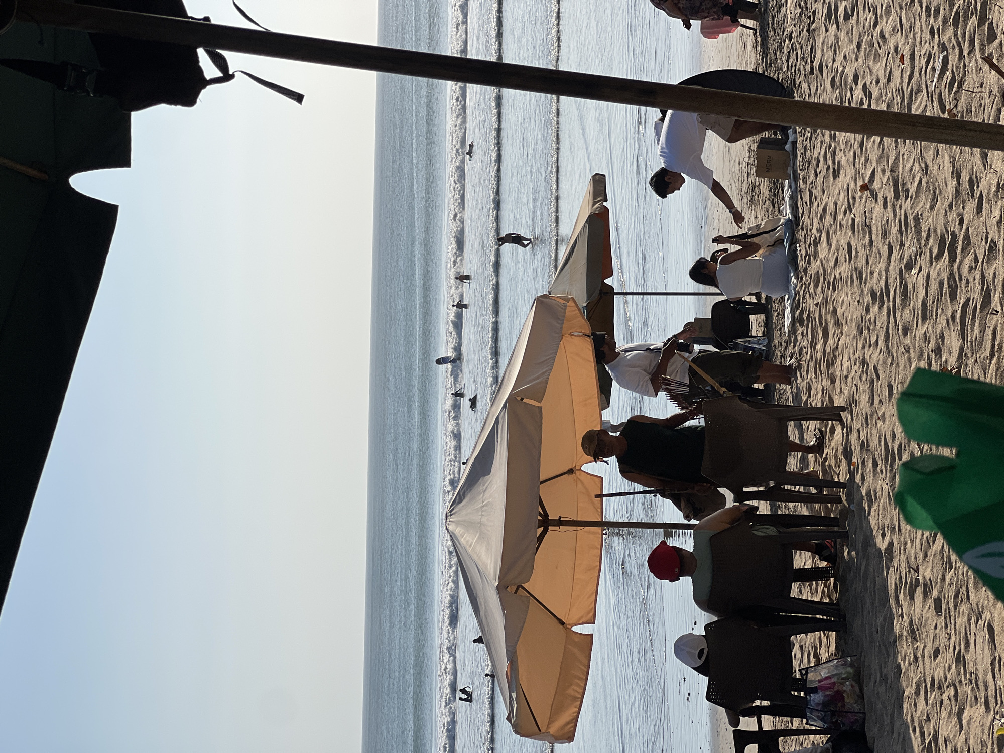 The image captures a lively scene on Kuta Beach, Indonesia, characterized by a bright, sunny day and gentle ocean waves. In the foreground, several people are relaxing on the sandy shore under the shade of beach umbrellas. Two individuals are seated in chairs, facing away from the camera, one wearing a red cap and the other a white cap. To their right, a person in a dark green sleeveless shirt and a cap stands near a beach umbrella, appearing to be interacting with another person. Further to the right, a man in a white shirt and shorts leans down to hand something to a woman sitting on a blanket on the sand. The ocean in the background is dotted with several surfers riding the waves, creating a dynamic and recreational atmosphere. The clear blue sky and the calm sea suggest a pleasant day at this popular Indonesian tourist destination.