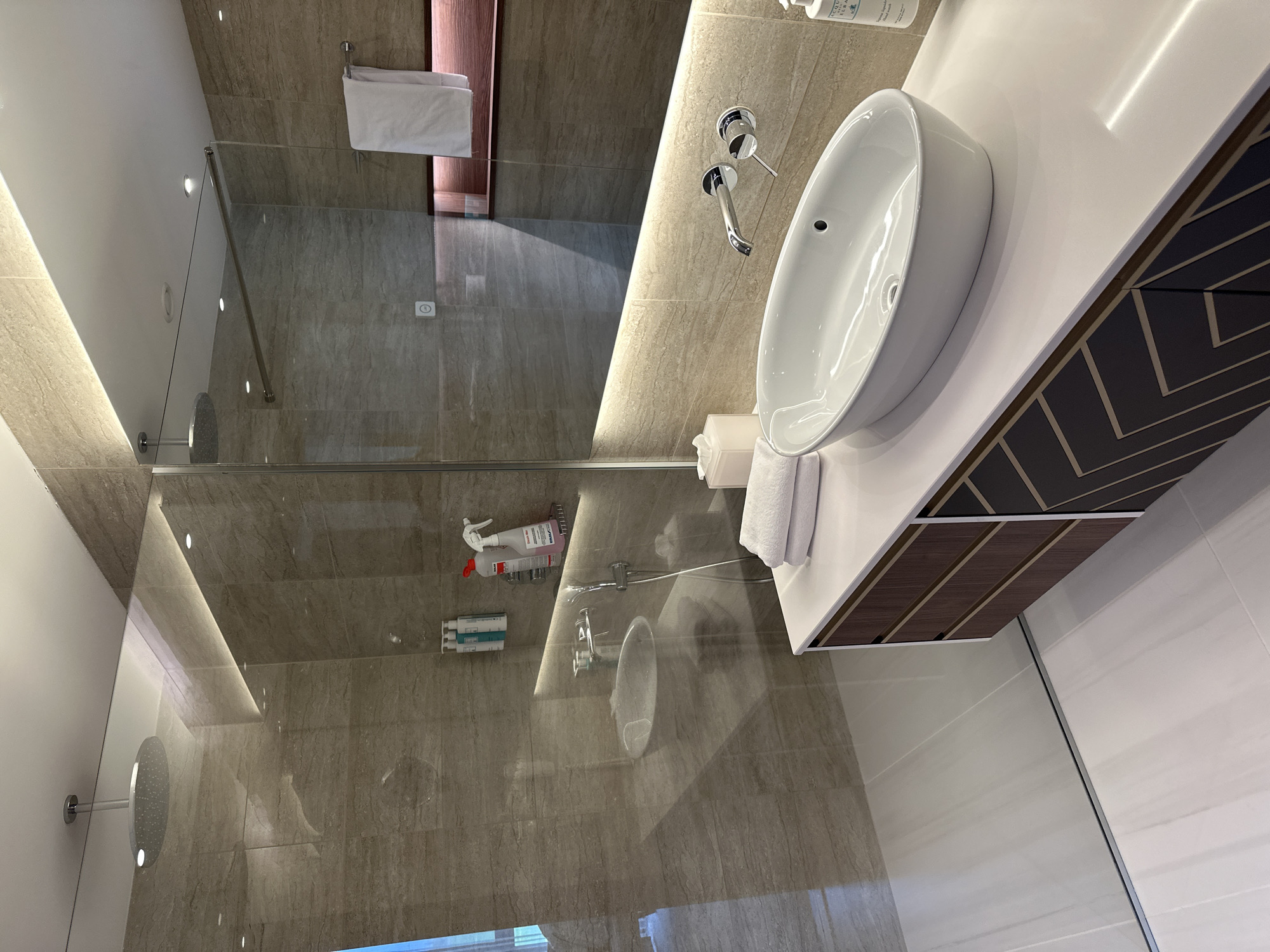 This is a high-angle, indoor shot of a bathroom in Kazanlak, Bulgaria. The main elements are a sink, a shower, and the surrounding bathroom fixtures.
The sink is a modern, white, oval-shaped basin sitting atop a white countertop. The cabinet below the counter is decorated with a herringbone pattern of alternating dark and light wood. A chrome faucet is mounted on the wall behind the sink.
The shower is enclosed by a glass partition and has a rainfall showerhead. A dispenser containing cleaning supplies is mounted on the wall. There is a white towel hanging on a rack and some toiletries on the shower shelf.
The walls of the bathroom are covered in neutral-colored tiles. The floor is also tiled, with lighter-colored tiles.