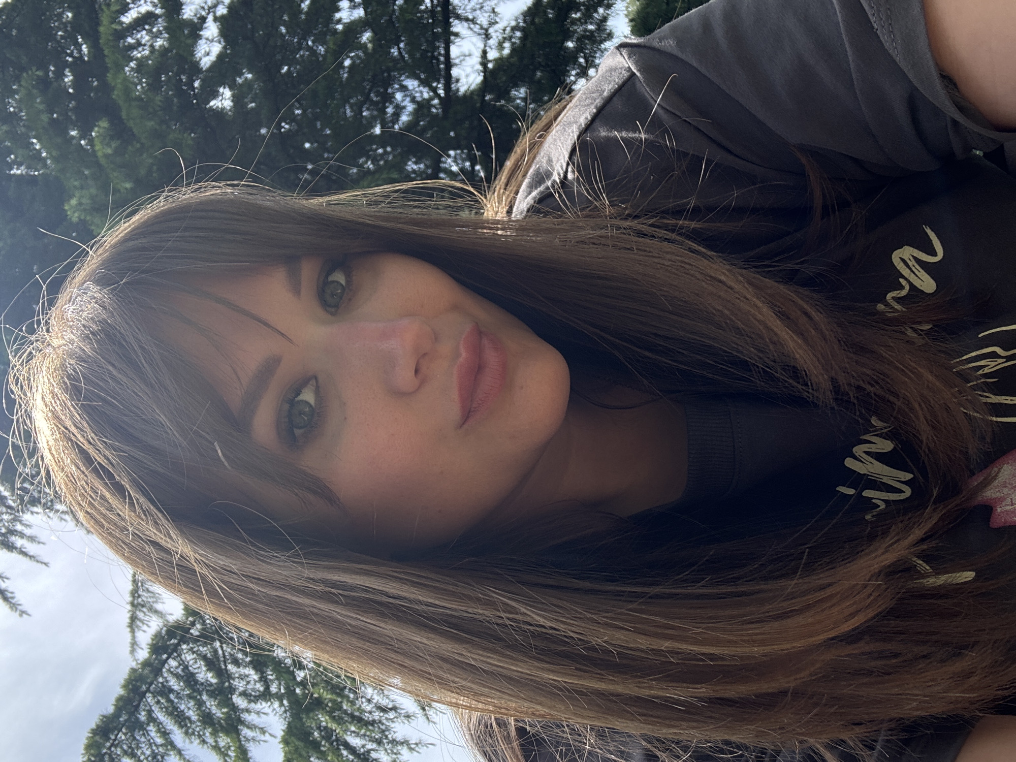 A selfie shows a fair-skinned woman with brown hair and bangs, wearing a dark-colored t-shirt, with trees visible in the background. The location is inferred to be in or near Гурзуфский поселковый совет, Ukraine, based on the user's request. The image is angled, taken from a higher perspective, showing the woman's face tilted towards the camera. The lighting is natural, with some areas of bright sunlight.
